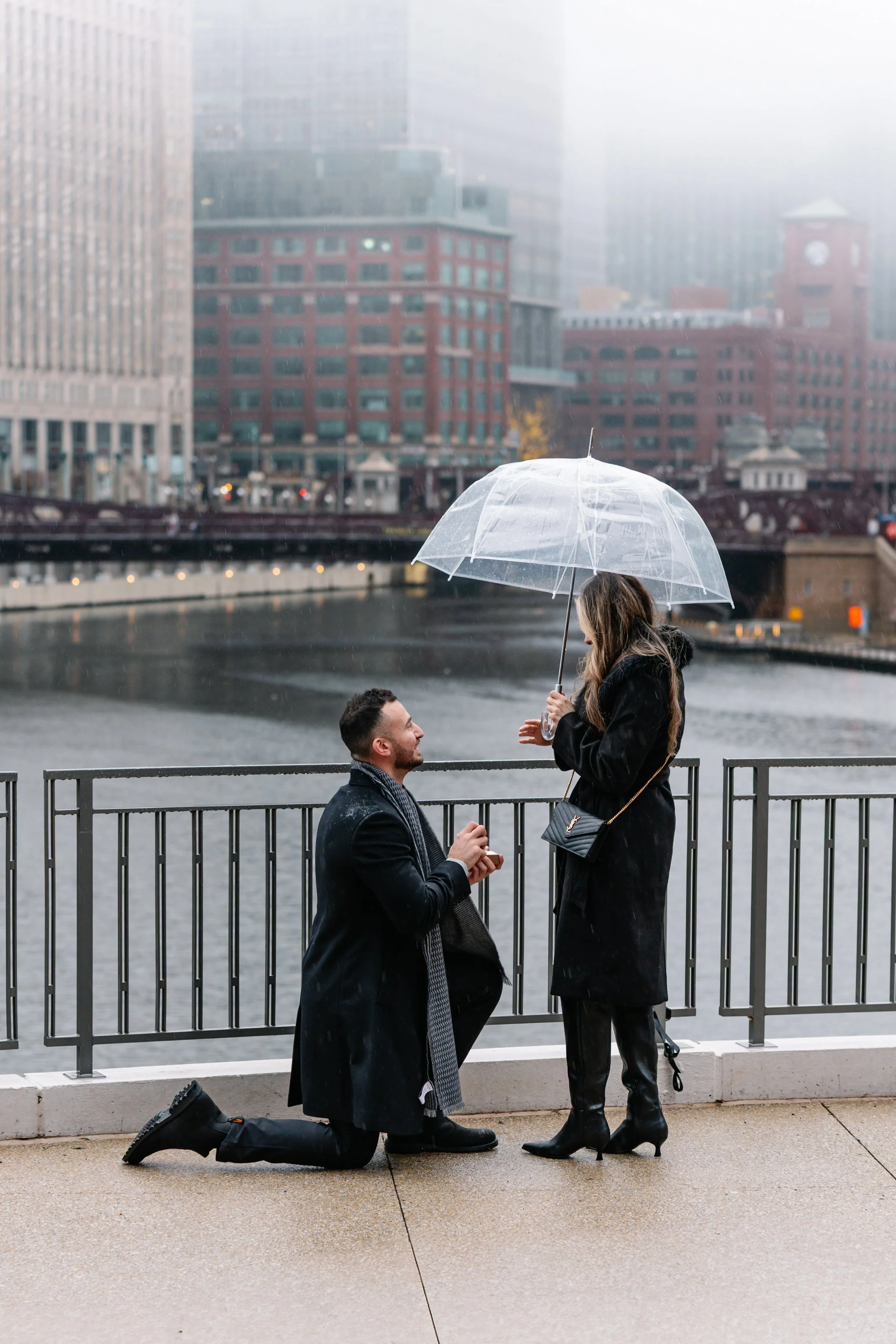 A woman proposes to a man by a river in a city on a rainy day, with a clear umbrella covering them.