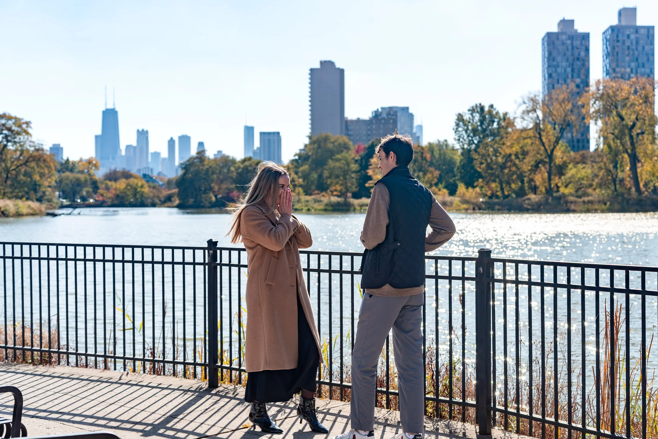 A man and woman stand by a railing near a lake in a city park during fall, with city skyscrapers in the background. The woman appears surprised, covering her mouth, while the man faces her with his hands on his hips.