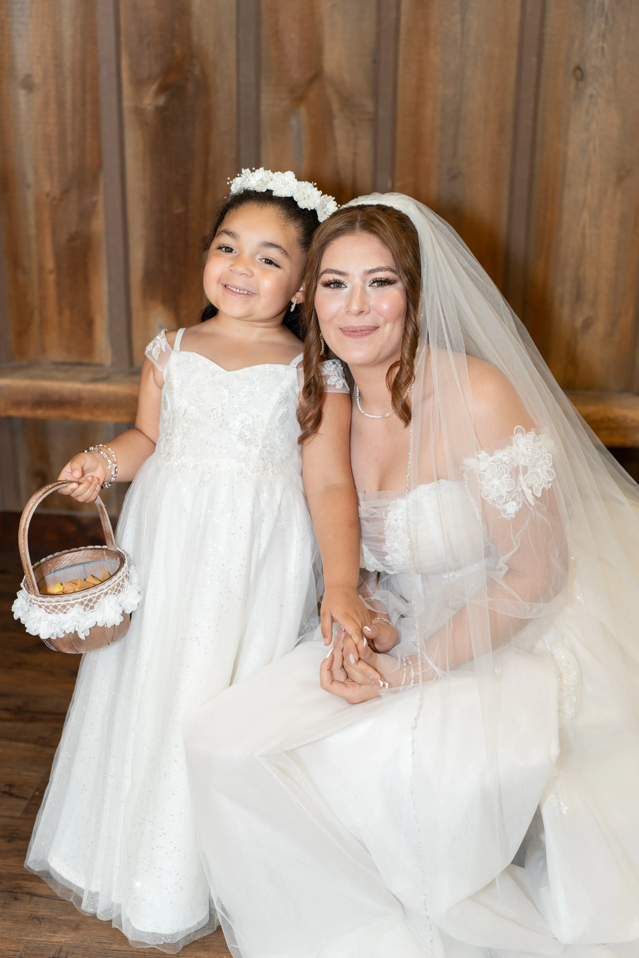 A bride kneeling next to a young girl dressed in a white dress, both smiling at the camera in front of a wooden backdrop.