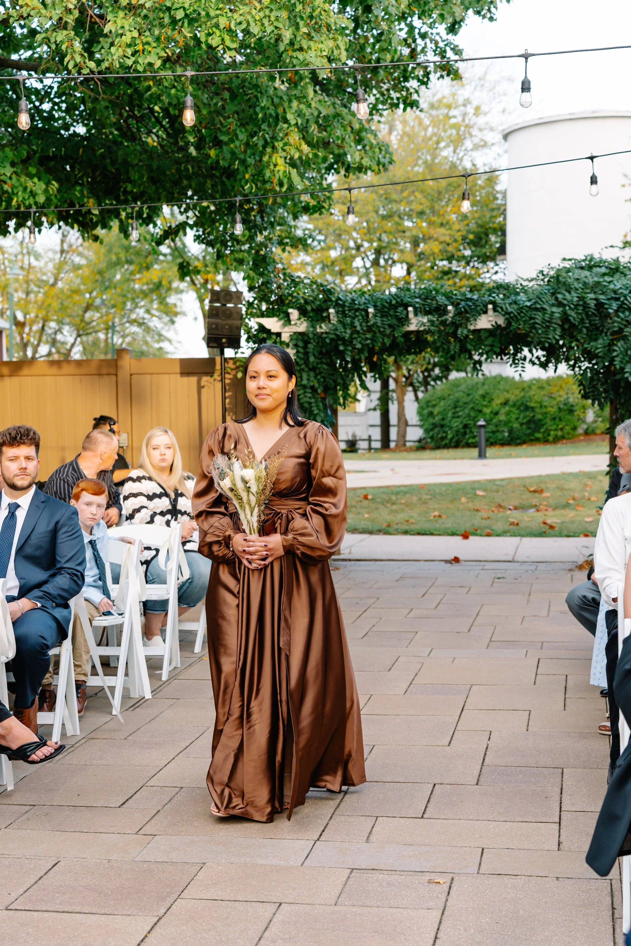 A woman in a long, brown satin dress holding a bouquet of flowers walking down an outdoor aisle during a wedding ceremony, with guests seated on either side and string lights hanging overhead.