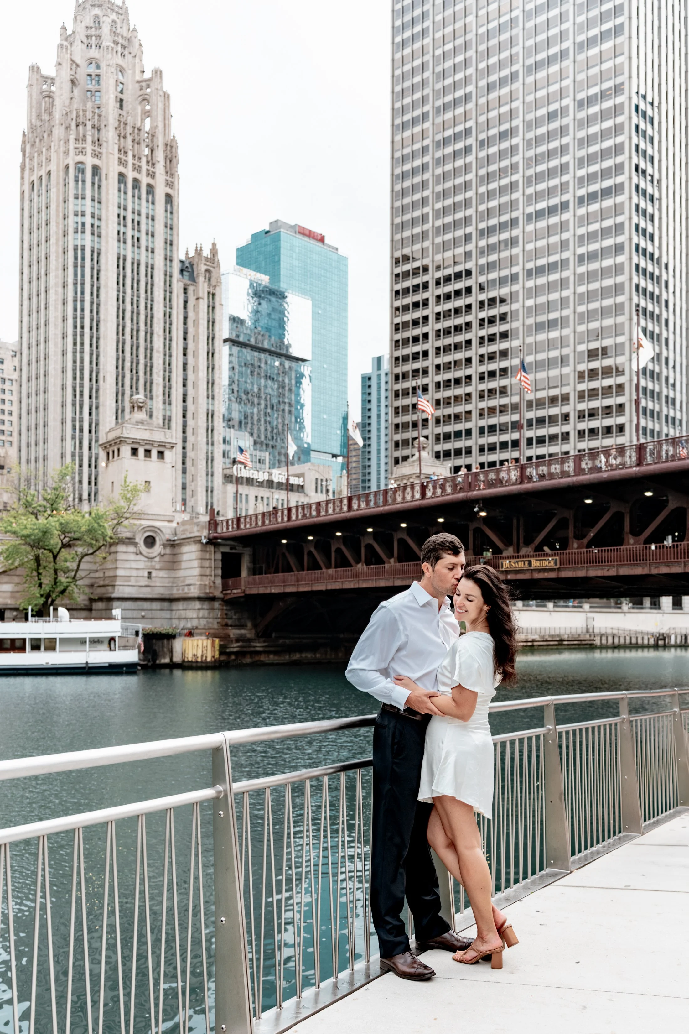 A couple standing close together by a river in downtown Chicago, with the Chicago River and city buildings, including the Wrigley Building and Tribune Tower, in the background.