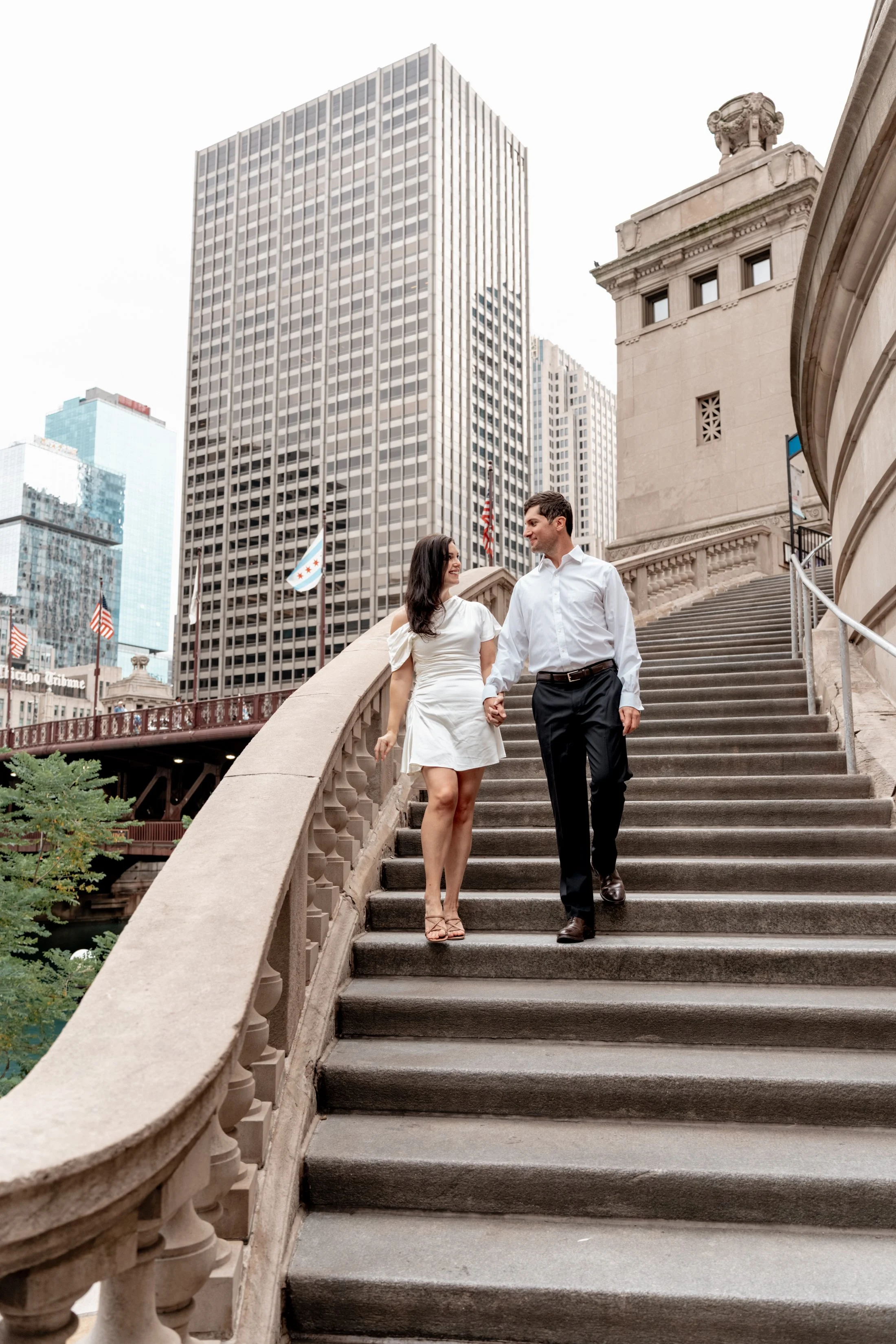 A couple walking down the stairs of a historic building in a city, holding hands and smiling at each other, with tall modern buildings and American flags in the background.