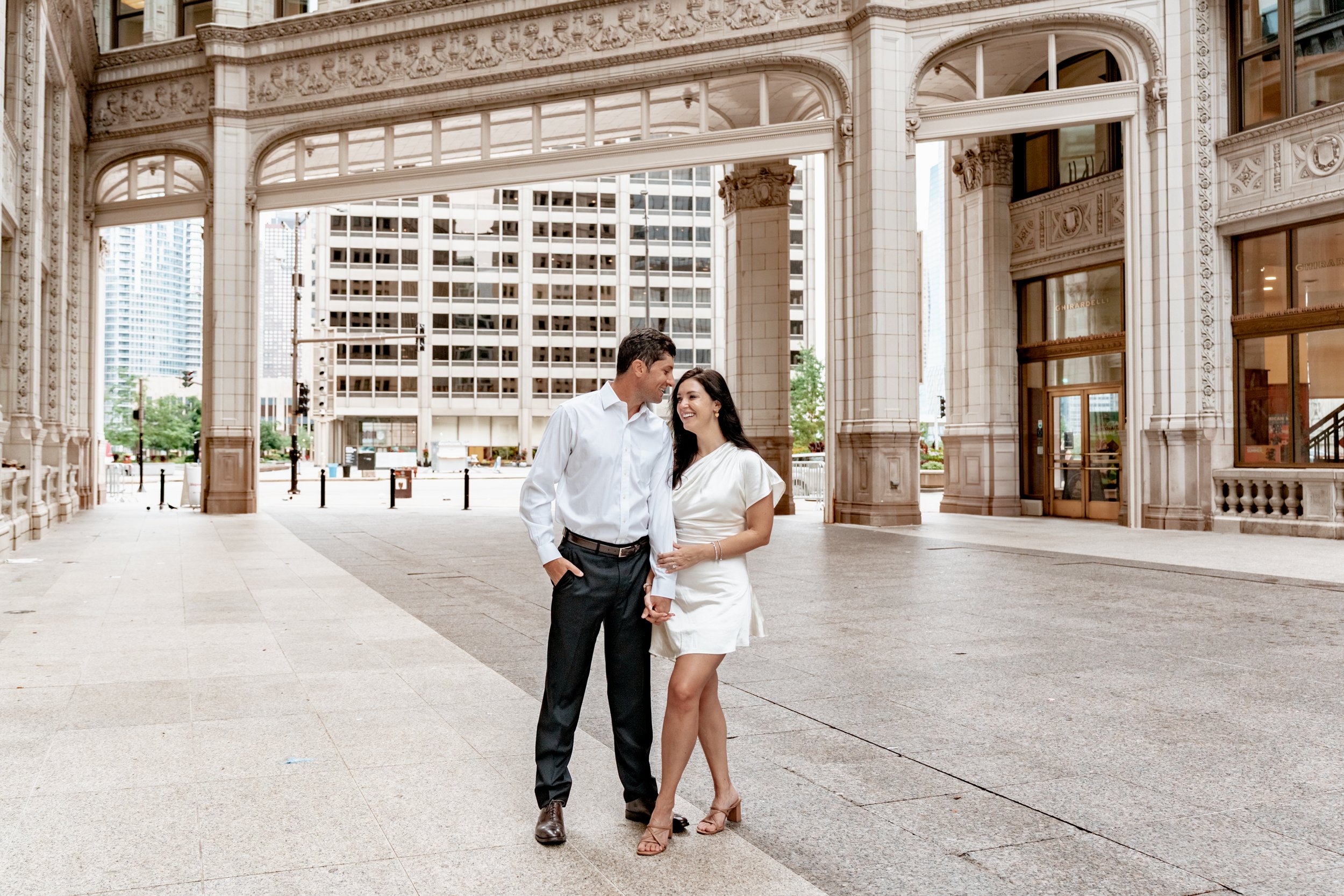 A smiling man and woman standing close and holding hands in an urban area with ornate architectural buildings and modern skyscrapers in the background.
