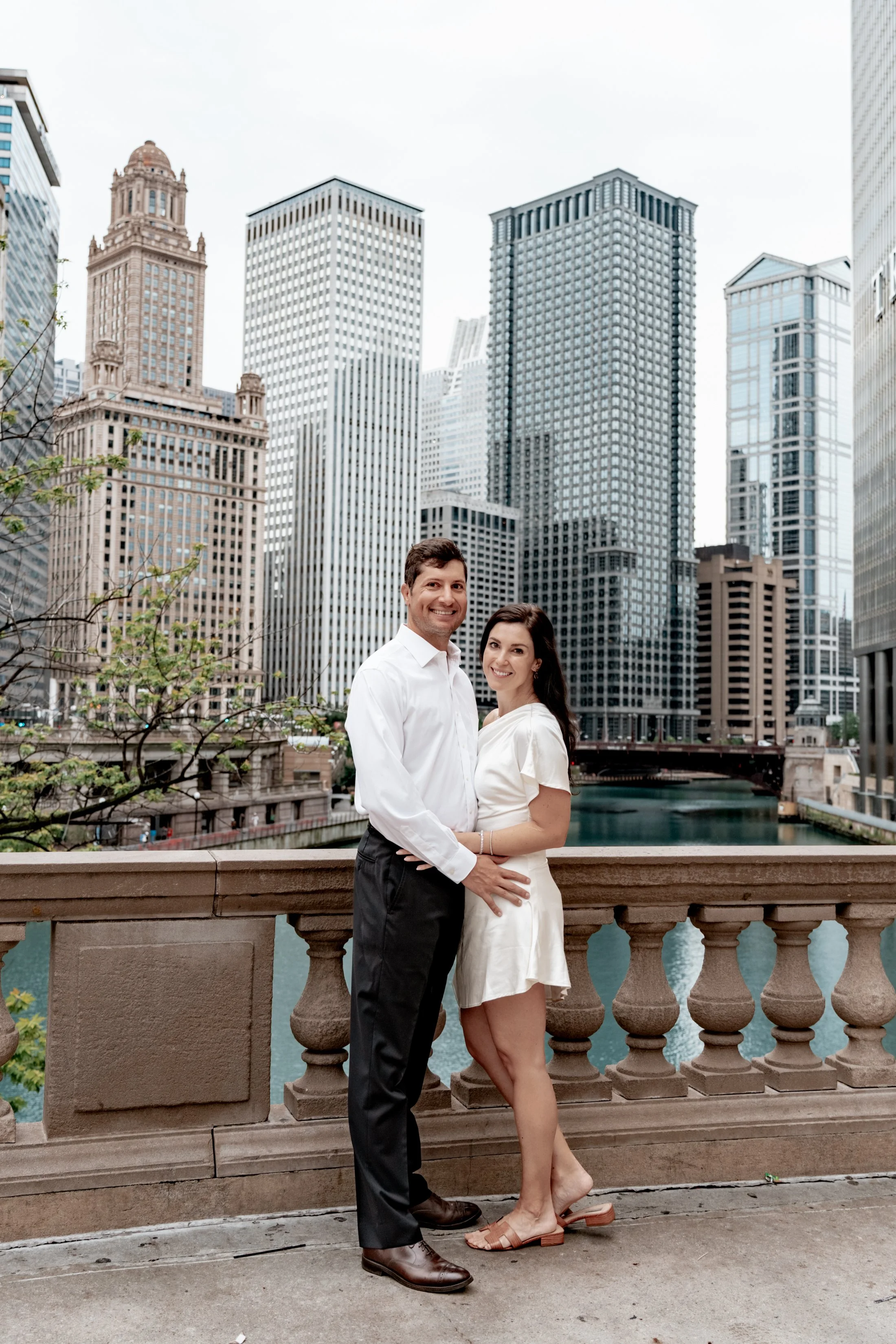 A smiling couple stands close together on a bridge with a cityscape of tall buildings and water in the background.