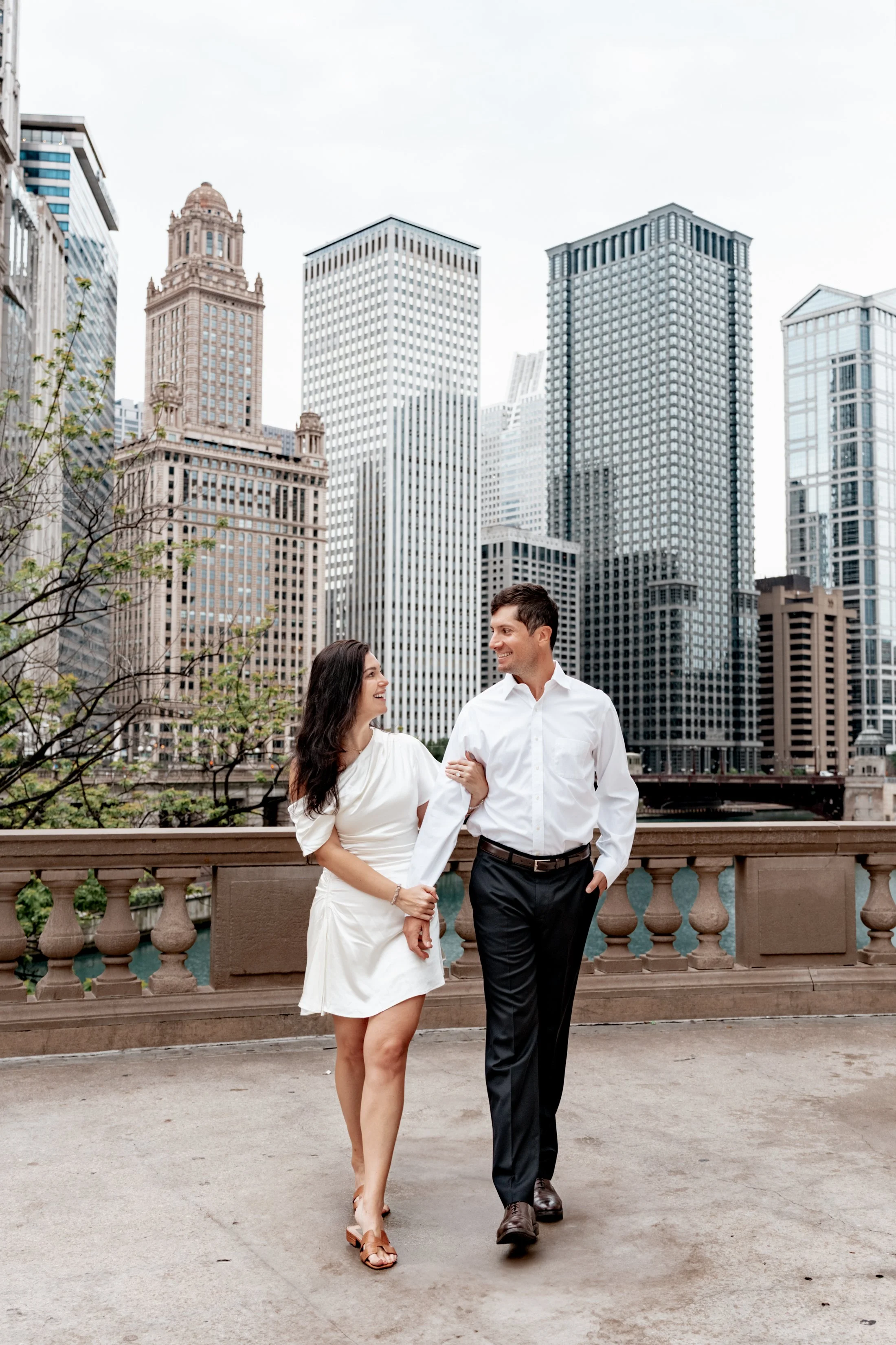 A couple walking arm-in-arm on a city balcony with tall skyscrapers in the background.