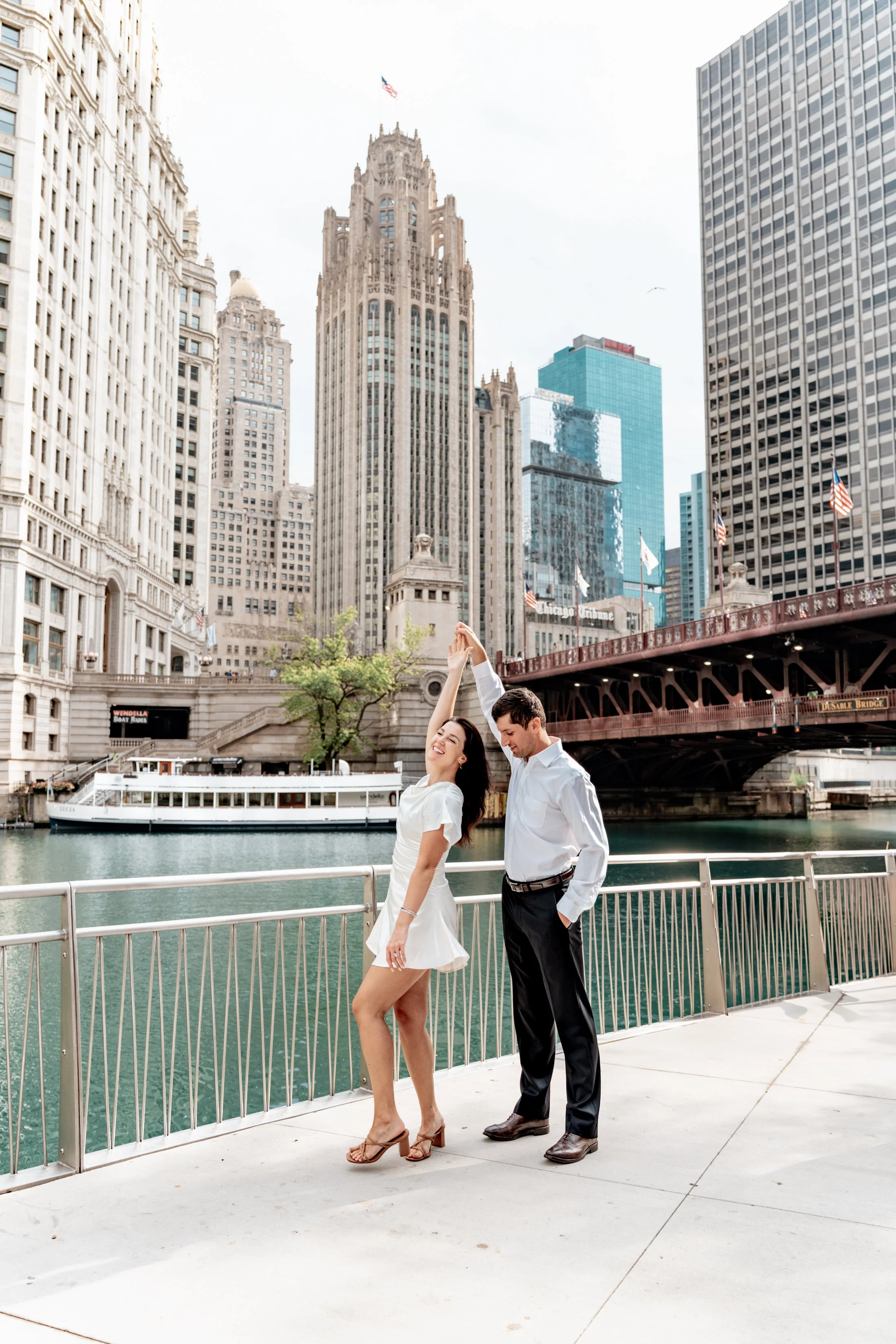 A couple dancing by the Chicago River in front of downtown skyscrapers and a bridge.
