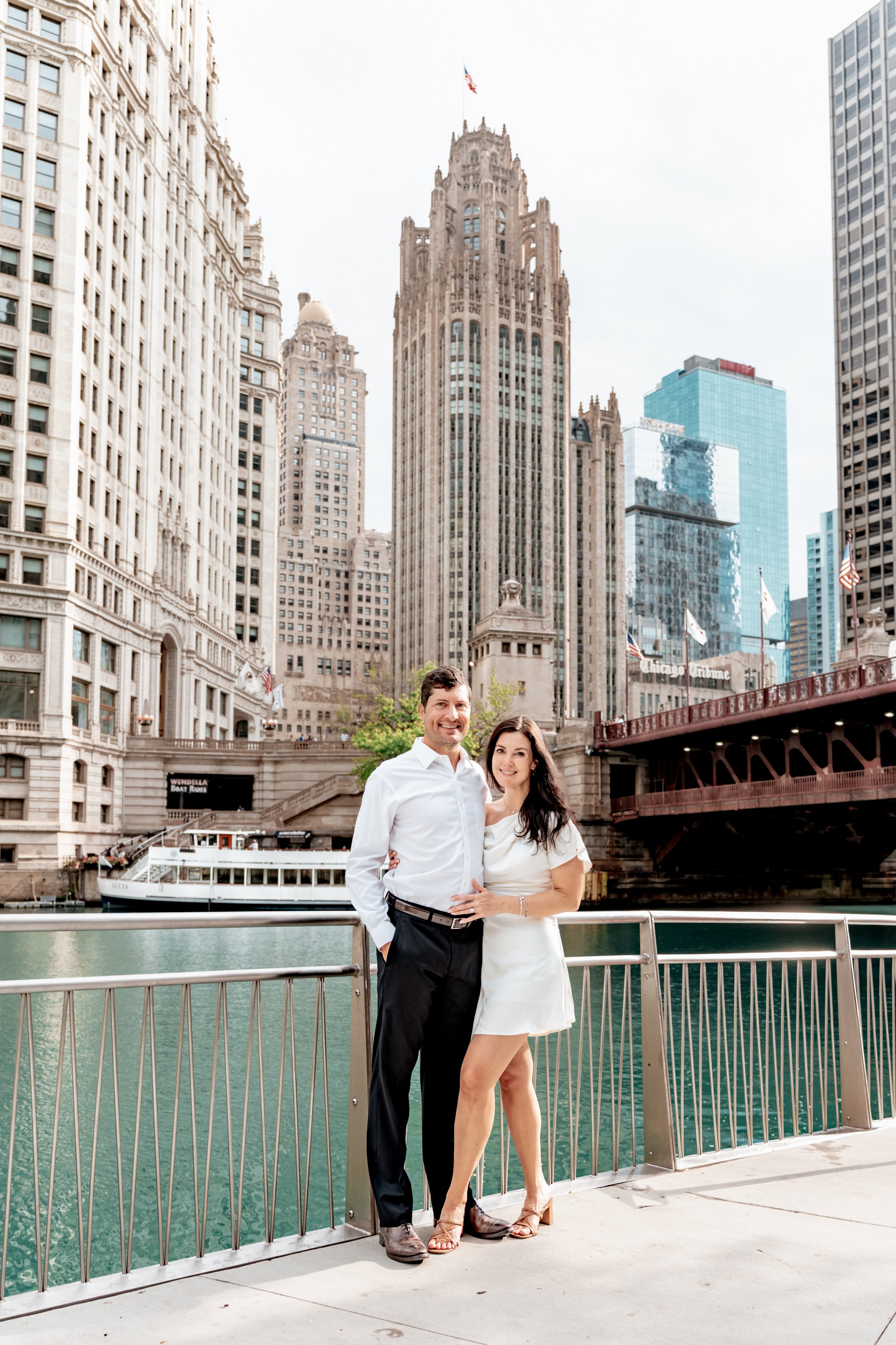 A couple standing on a riverwalk promenade in Chicago with tall skyscrapers in the background, including the Wrigley Building and Tribune Tower.