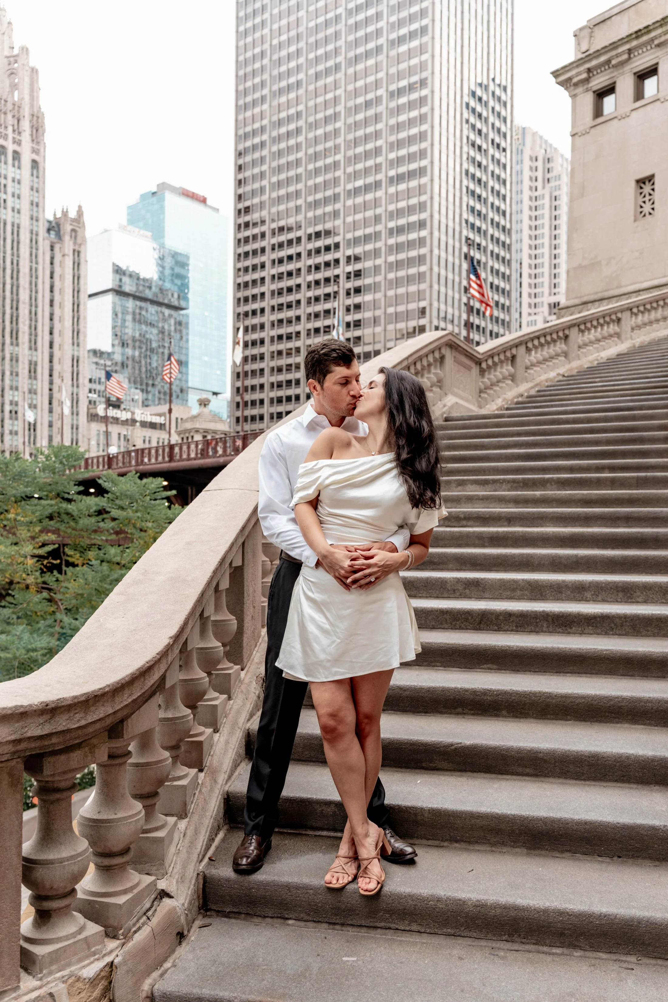 A couple sharing a kiss on a staircase in an urban setting, with skyscrapers and American flags in the background.