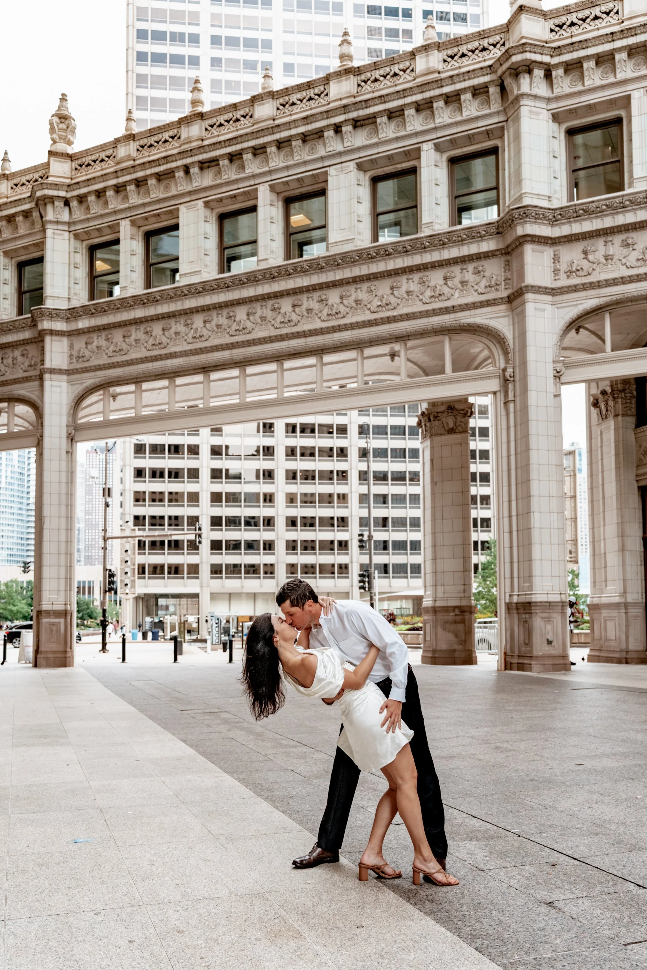 A couple dancing and kissing under an ornate city archway surrounded by tall buildings.