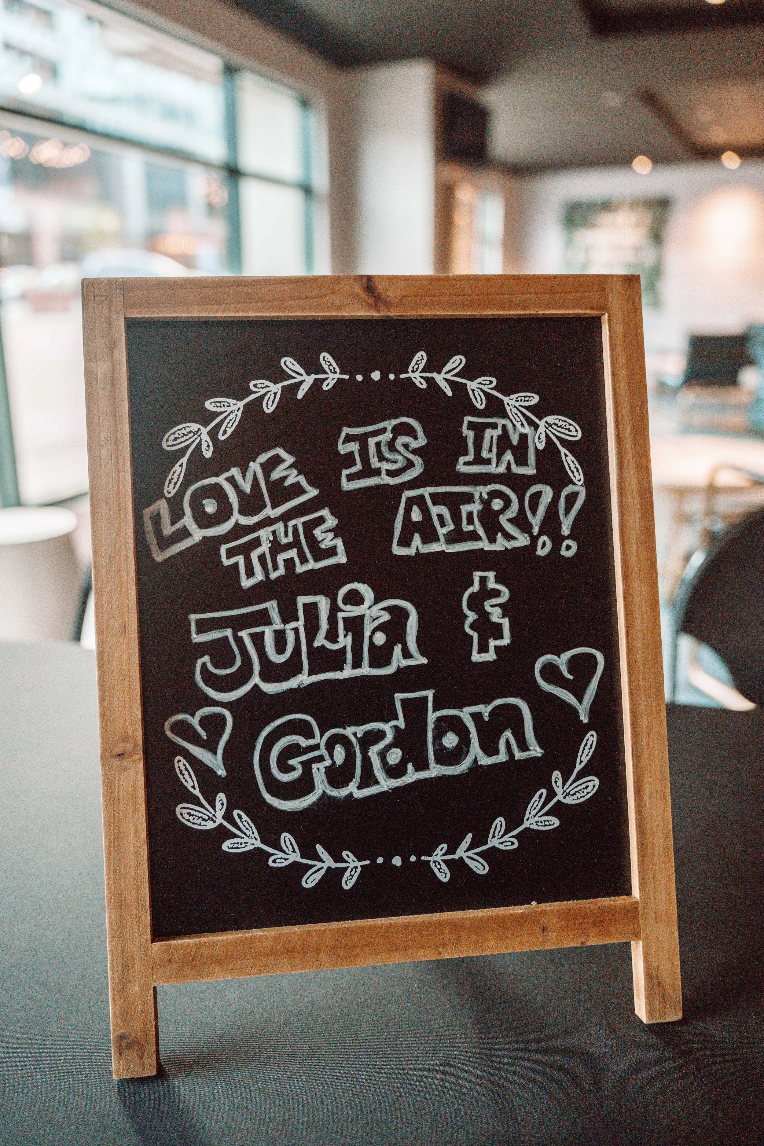 A chalkboard sign with decorative text that reads "Love is in the air!!" and mentions a wedding between Julia and Gordon, surrounded by a leafy border and hearts, inside a cozy cafe or restaurant.