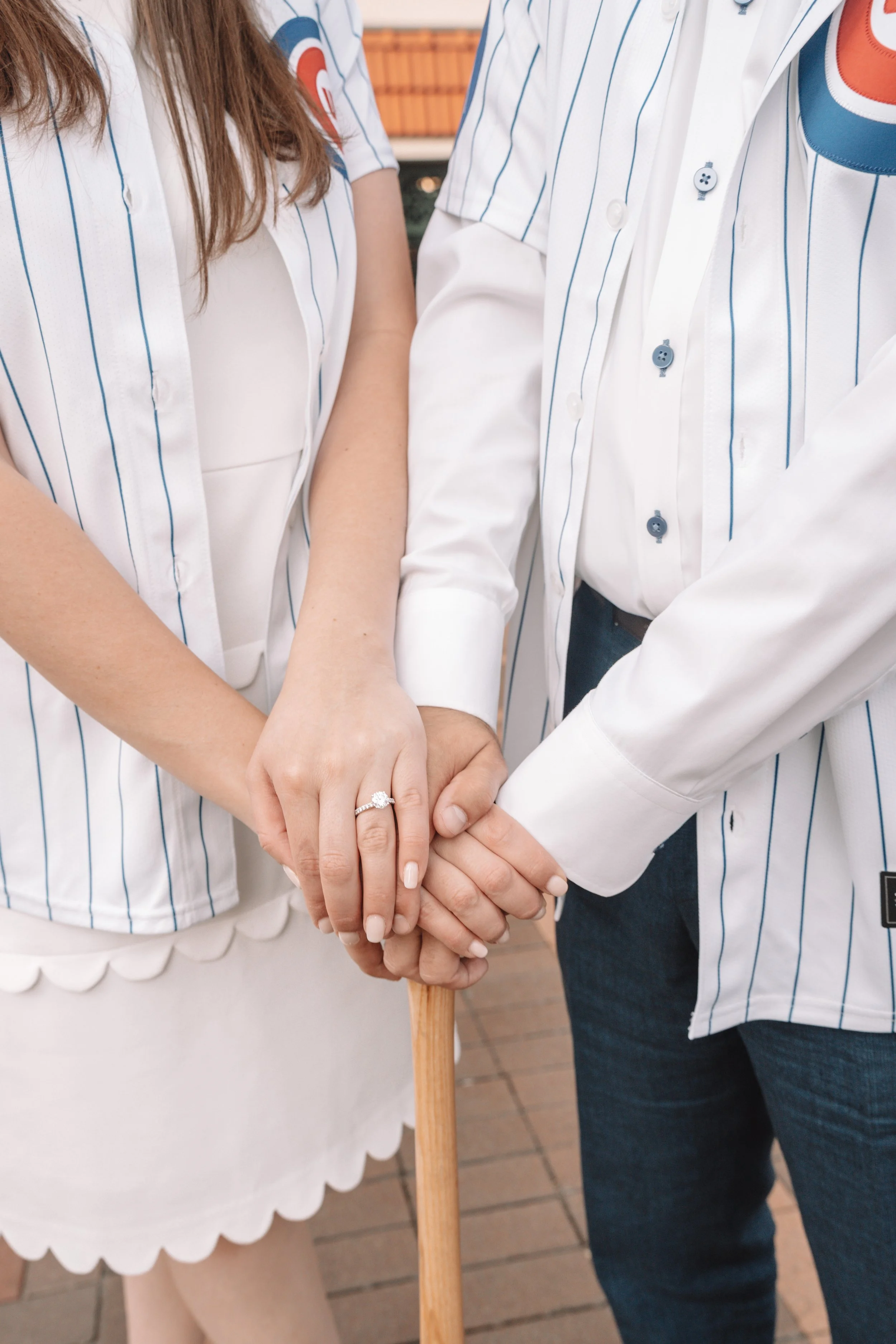 A couple holding hands, showing an engagement ring, with a cane visible in front of them. They are wearing matching white shirts with blue stripes.