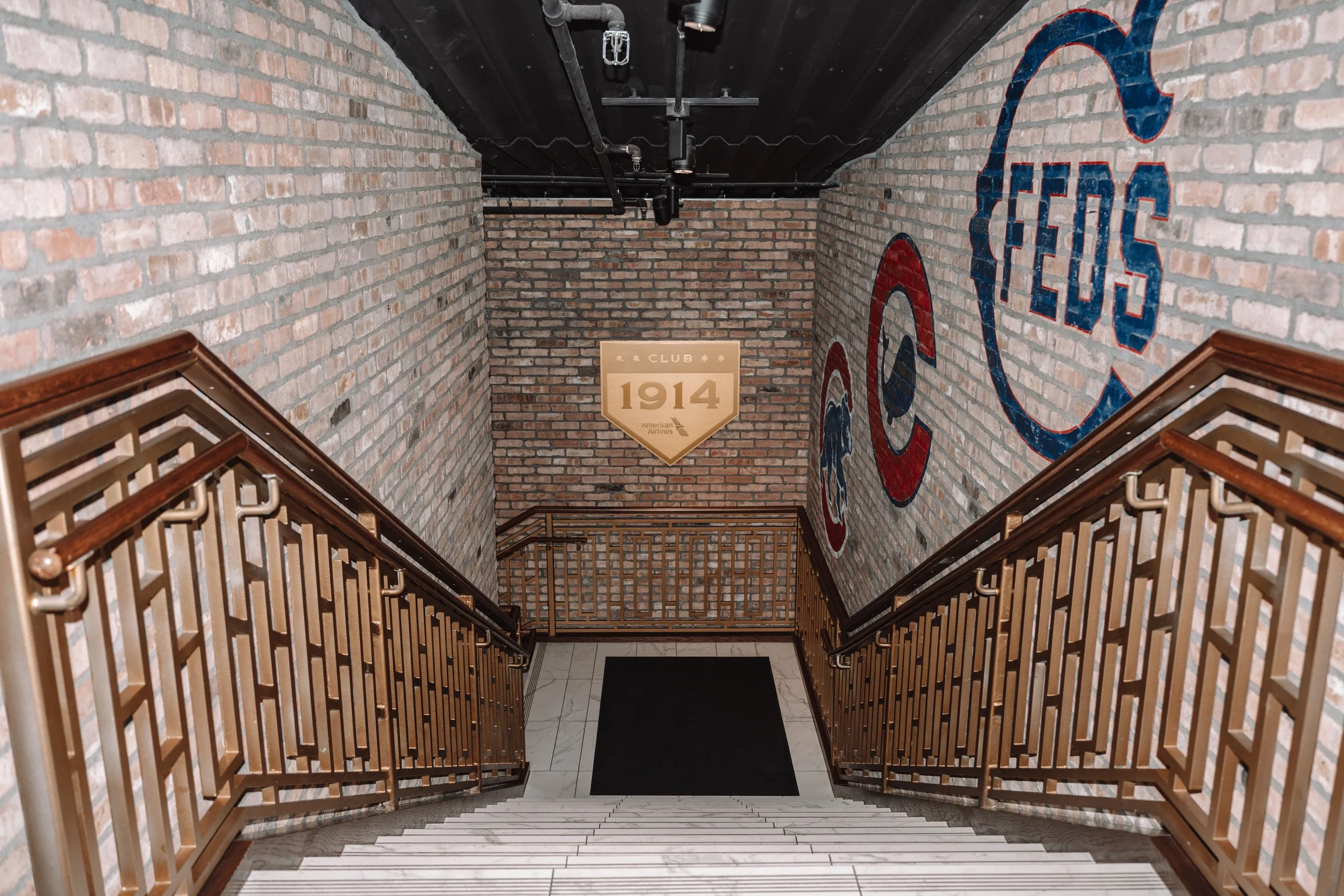 View of a staircase leading down to a hallway with exposed brick walls decorated with large logos of the Chicago Cubs and a sign reading "Club 1914" on the far wall.
