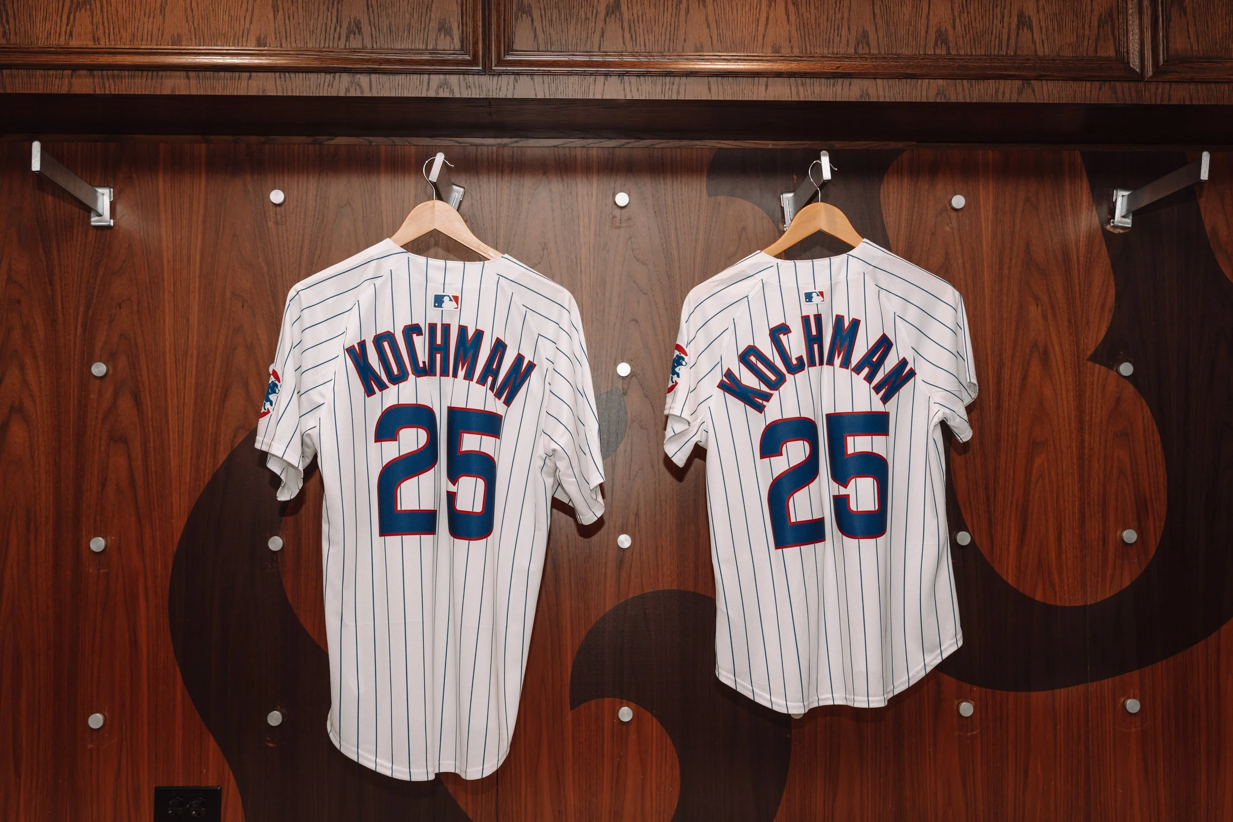 Two New York Mets baseball jerseys with the name 'KOCHMAN' and number '25' hanging on hooks in a locker room with wooden walls.