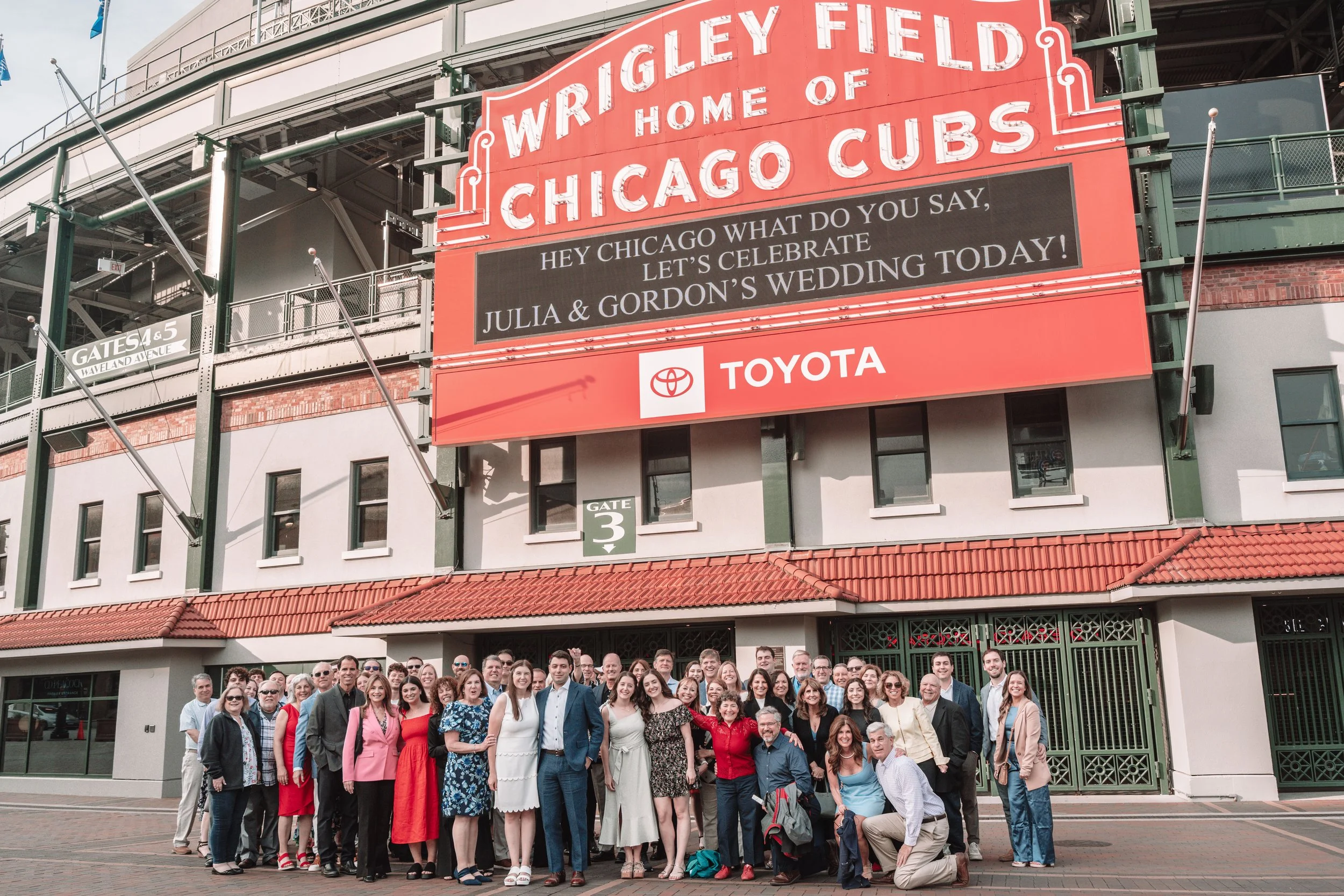 A large group of people posing in front of Wrigley Field, home of the Chicago Cubs, with a digital scoreboard displaying a message celebrating Julia and Gordon's wedding.