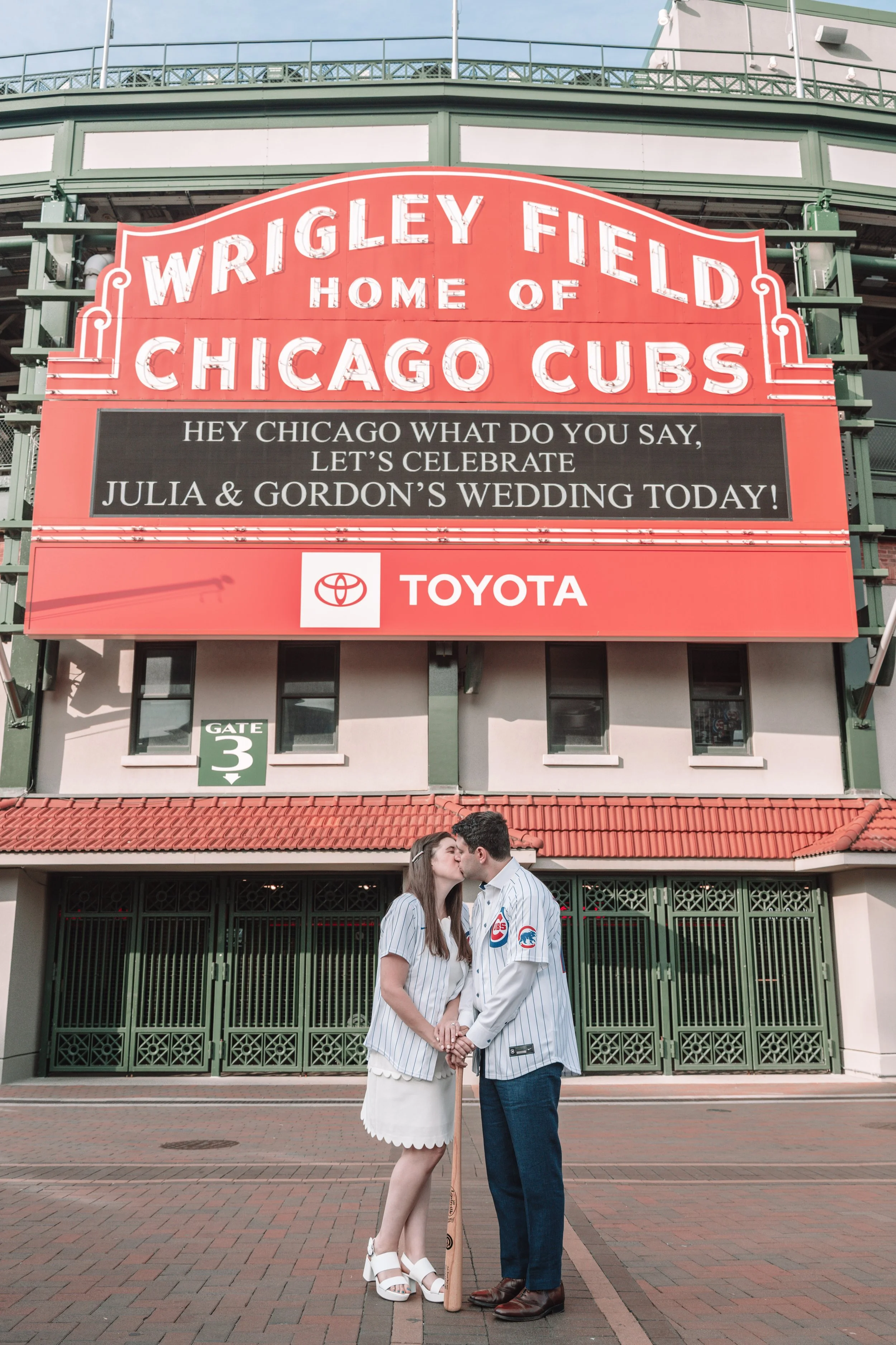 A couple dressed in Chicago Cubs baseball jerseys share a kiss in front of Wrigley Field in Chicago, with large signage announcing the stadium as home of the Chicago Cubs and a message celebrating a wedding.