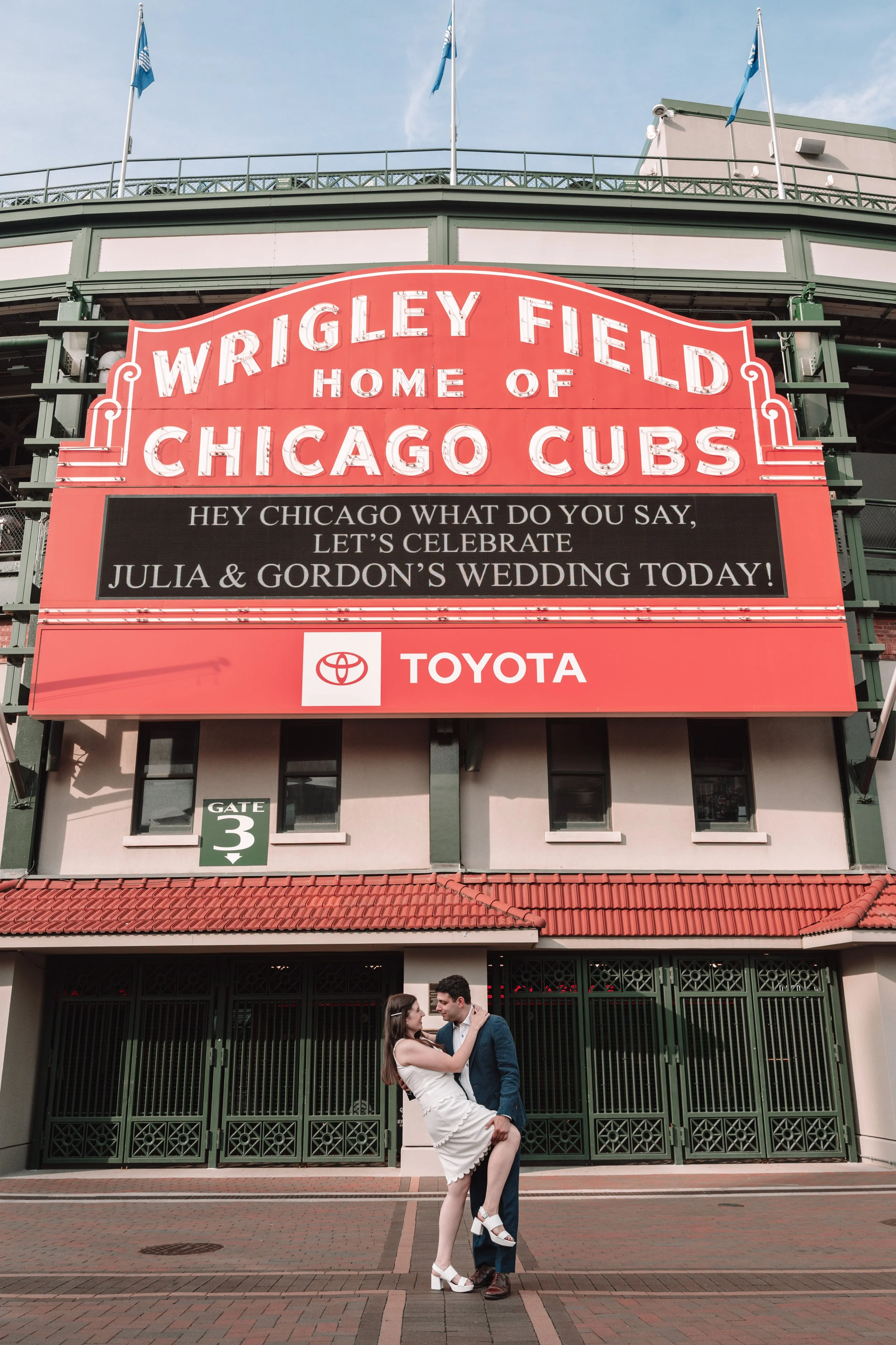 A couple is dancing in front of Wrigley Field, a baseball stadium in Chicago, with a large sign above them that reads "Wrigley Field Home of Chicago Cubs" and a message celebrating Julia and Gordon’s wedding.