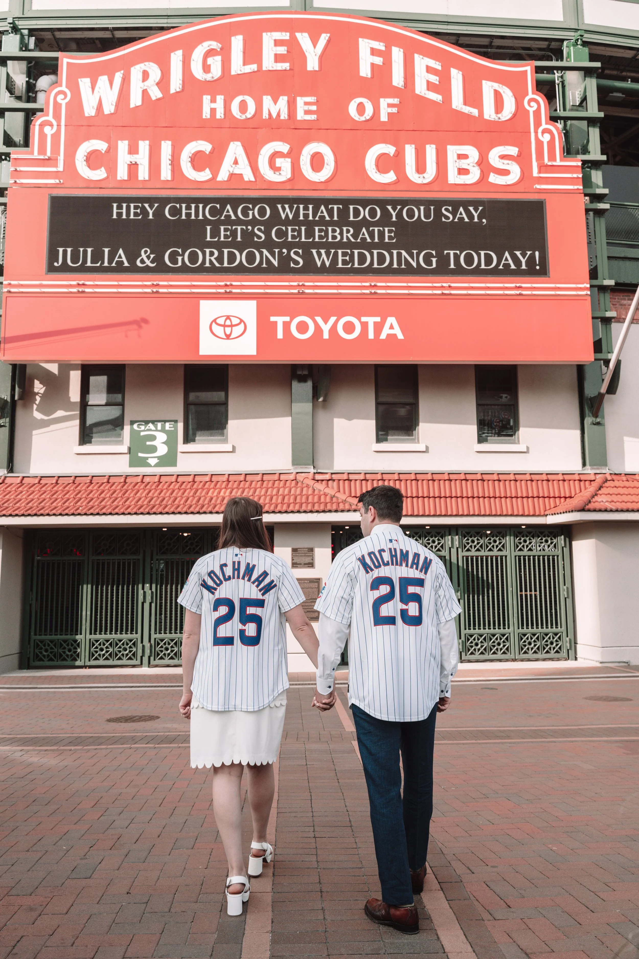 A man and woman holding hands, walking toward a baseball stadium entrance, both wearing Chicago Cubs jerseys with the number 25. The stadium sign above them reads Wrigley Field, home of the Chicago Cubs.