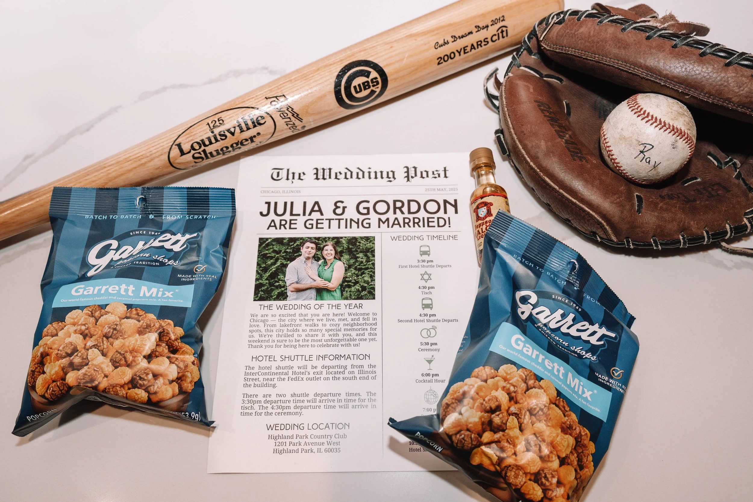 A baseball glove with a baseball inside, a wooden Chicago Cubs bat, two bags of Garrett Popcorn mixed flavor, a wedding invitation for Julia and Gordon, a small bottle of flaming hot sauce, and a baseball glove with a baseball inside on a white surfa