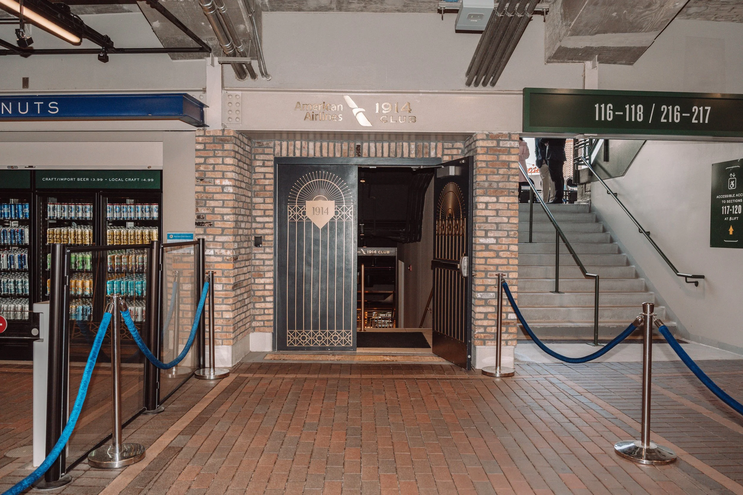 Entrance to the American Airlines 1914 Club located in an indoor space with brick walls, a black door with gold accents, and a staircase to the right. Blue velvet ropes cordon off the area.
