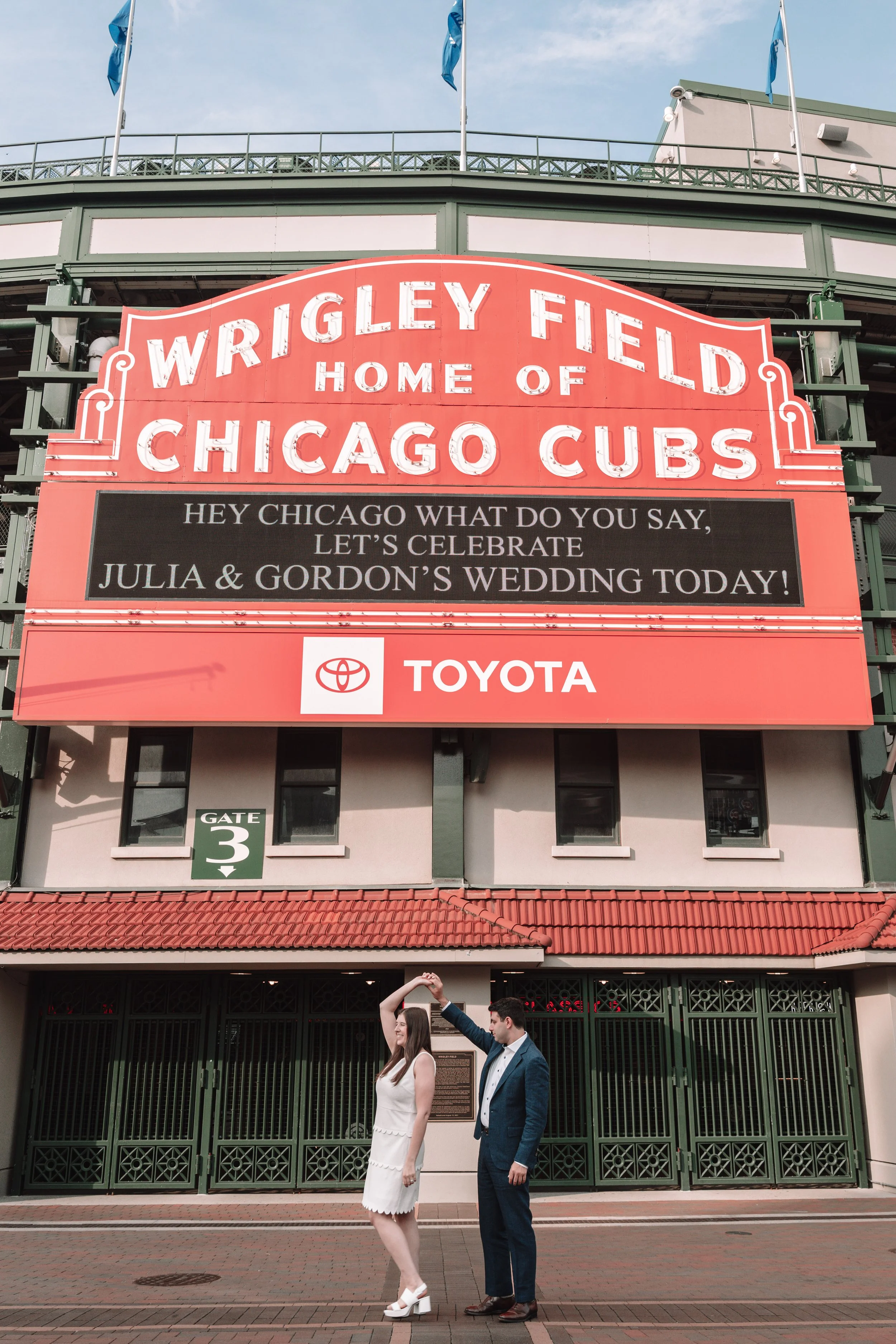 A couple is dancing in front of Wrigley Field, home of the Chicago Cubs. The woman is wearing a white dress and white heels, and the man is in a blue suit. Wrigley Field's signage and gates are visible in the background.