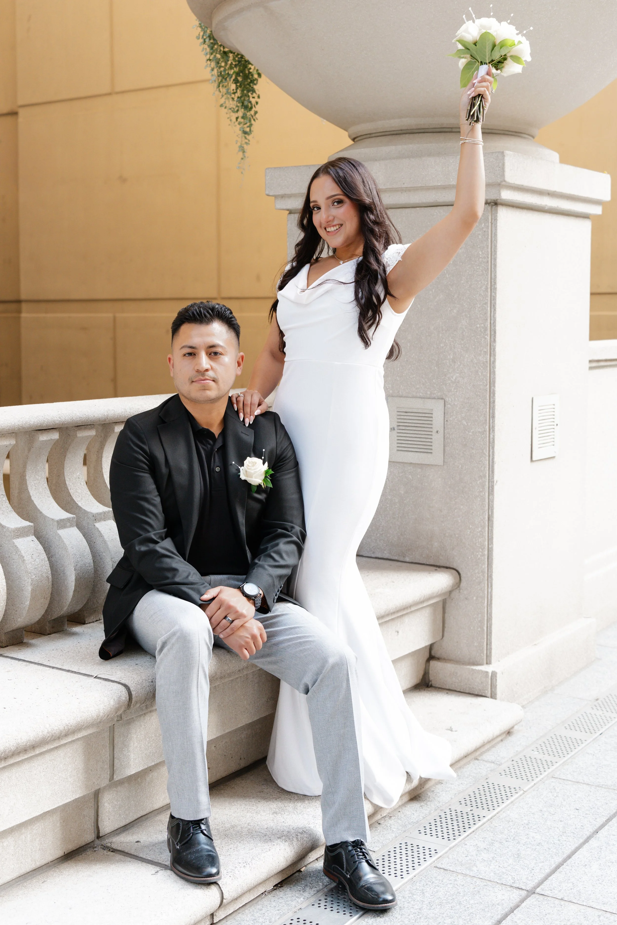 A woman in a white dress holding a bouquet of white flowers above her head and a man in a black blazer and gray pants sitting on a stone bench.
