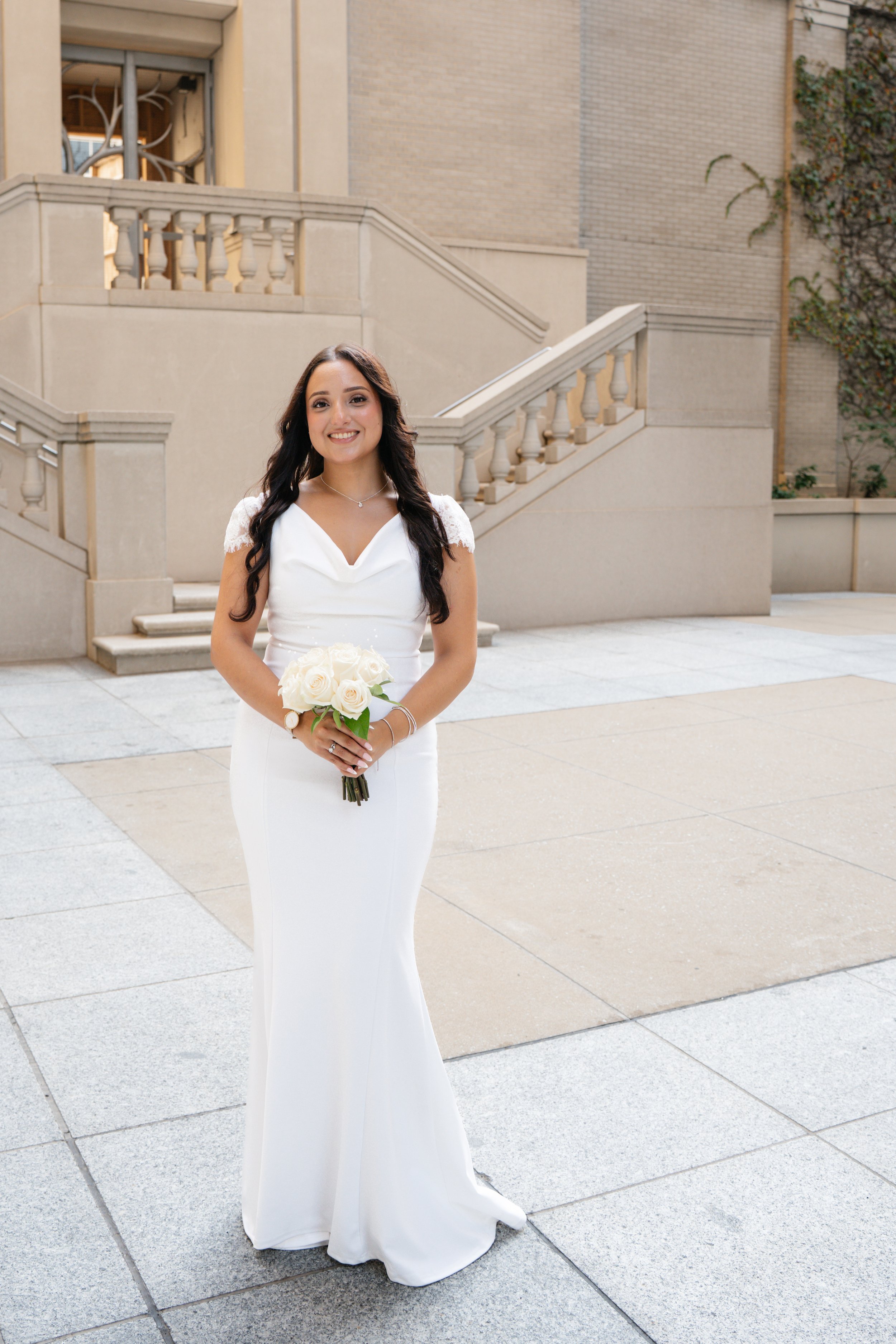 A young woman in a white wedding dress holding a bouquet of white roses, standing outdoors near beige stairs and walls, smiling at the camera.