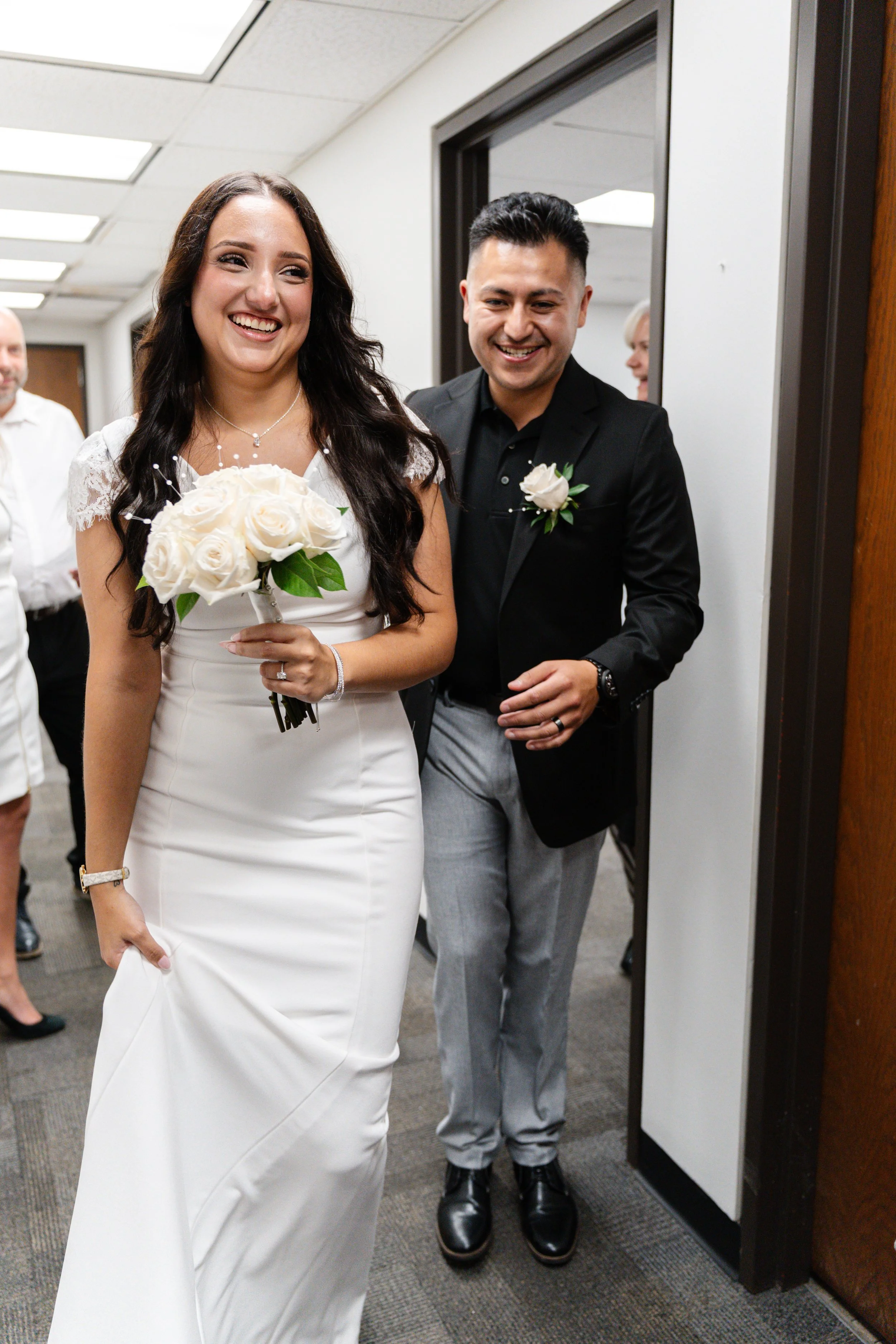 A bride in a white gown holding a bouquet of white roses and a groom in a black blazer with gray pants and a white rose boutonniere, smiling as they walk through a doorway with wedding guests in the background.