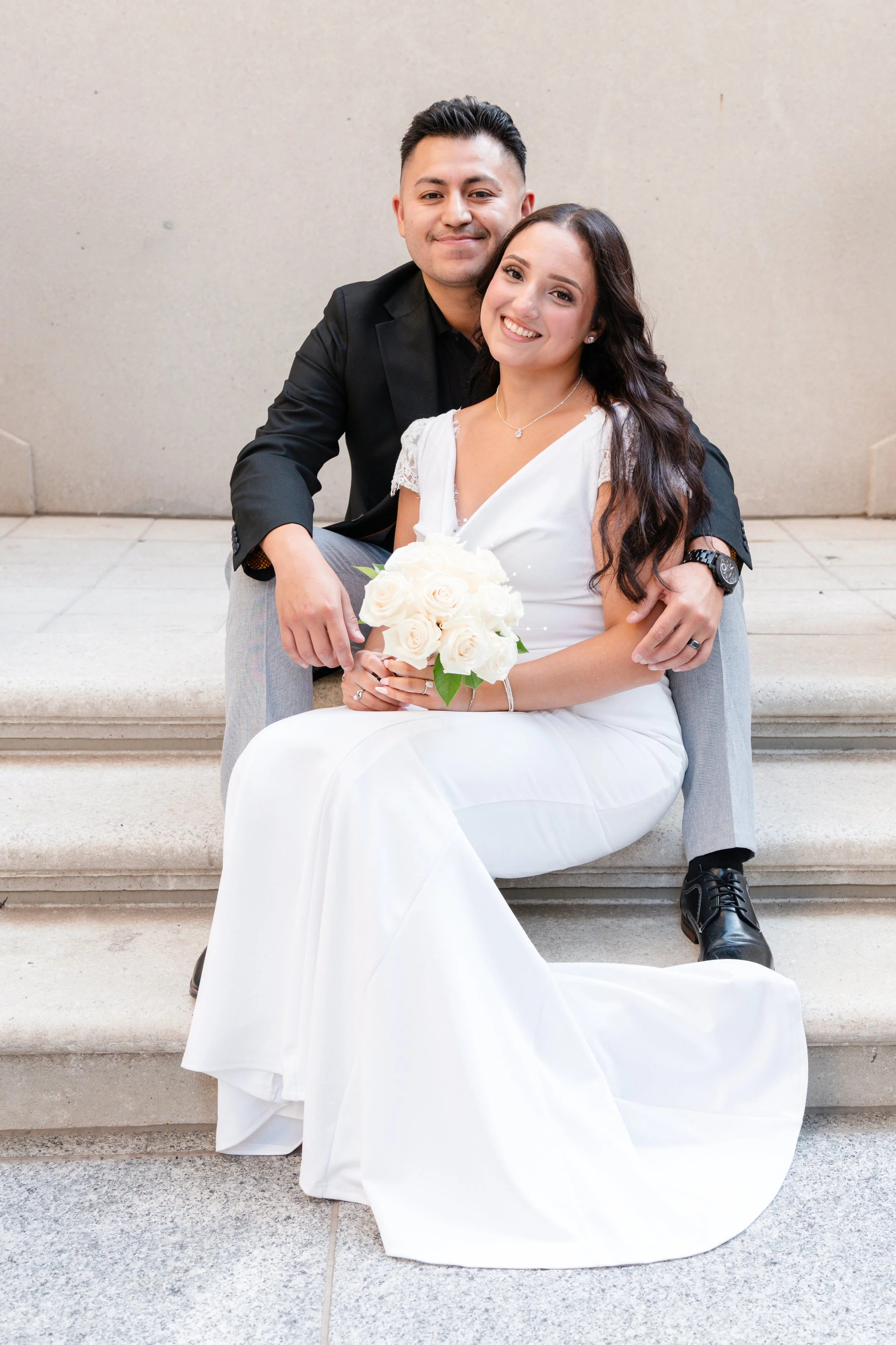 A smiling bride in a white wedding dress holding a bouquet of white roses, sitting on steps, with a groom in a black jacket and gray pants hugging her from behind.