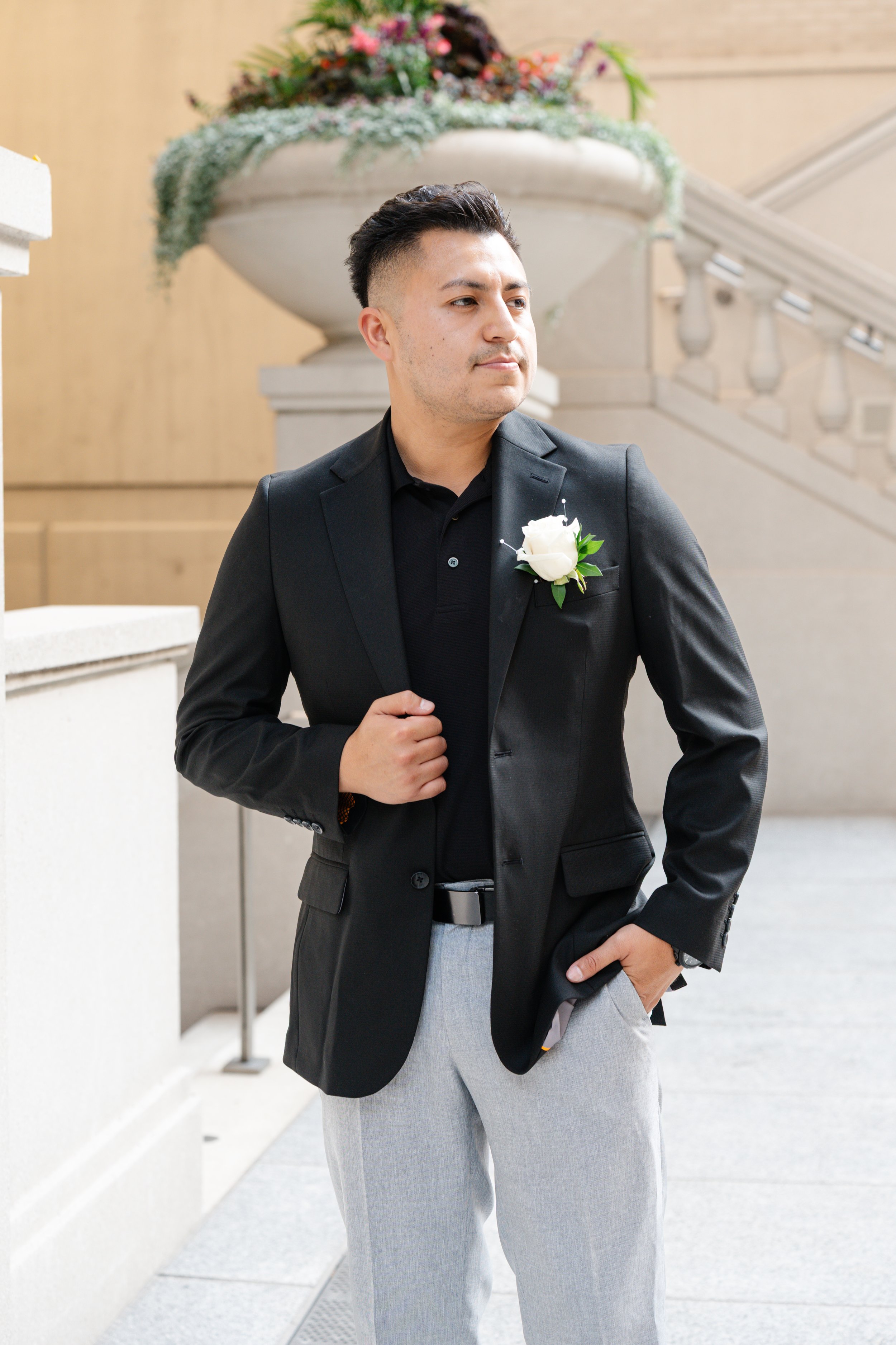 A man in a black blazer and light gray pants stands outdoors, with a large flower arrangement in the background. He has a white boutonniere pinned to his blazer and has his left hand in his pocket, looking to the side.