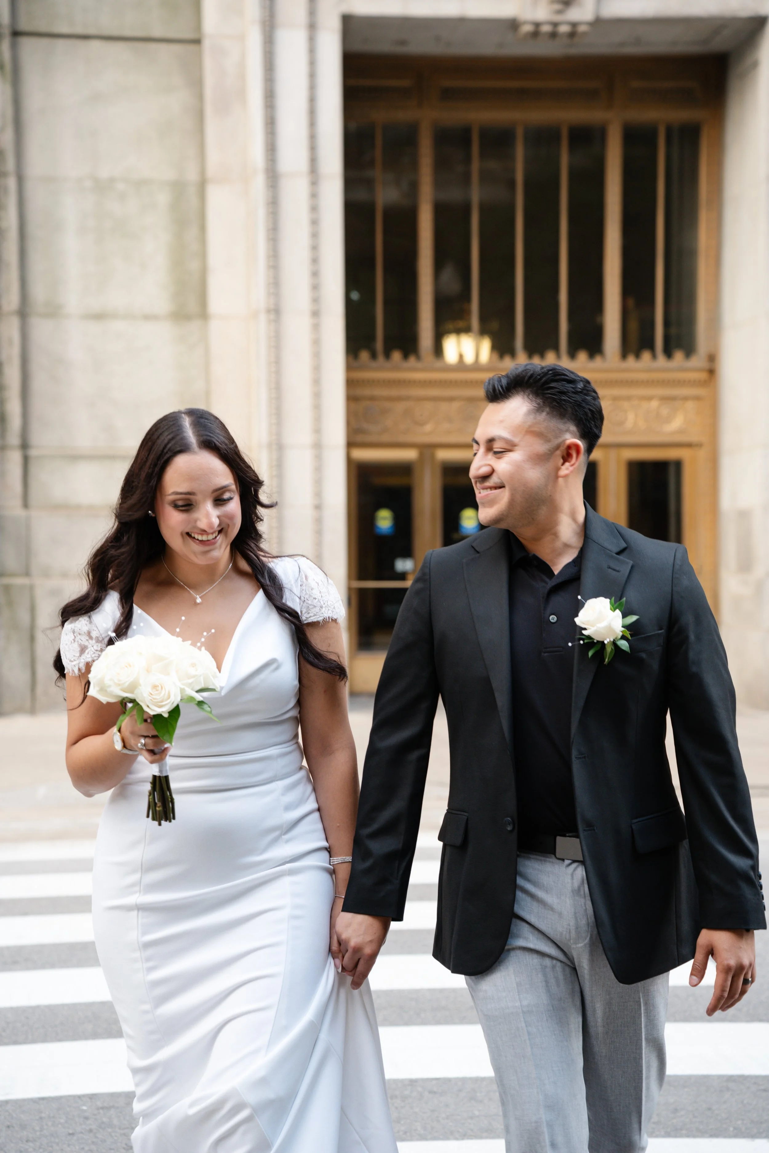 A couple in wedding attire walking hand-in-hand outside a building with large glass doors, smiling and holding a bouquet of flowers.