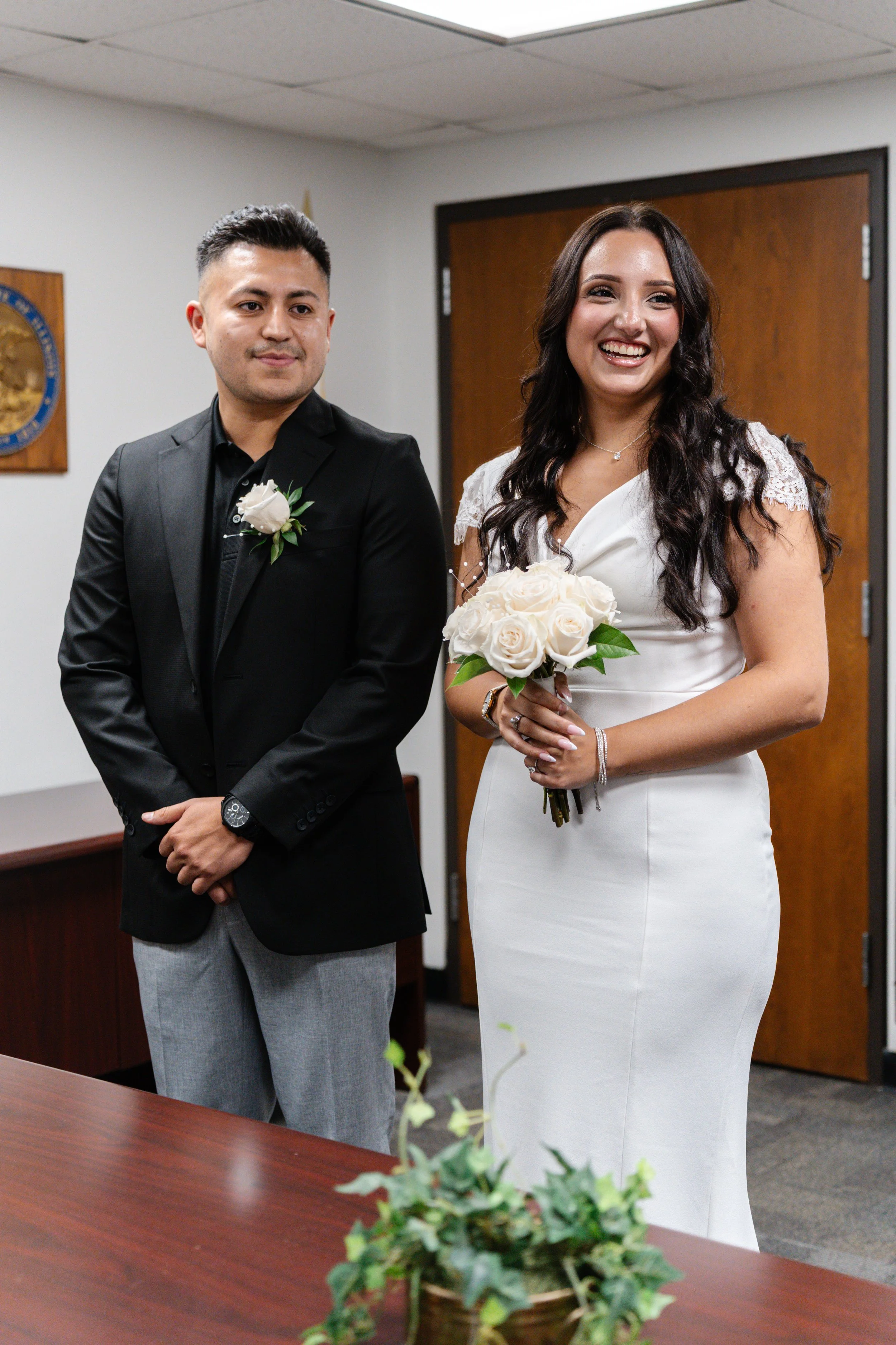 A bride in a white wedding dress holding a bouquet of white roses stands next to a groom in a black suit with a boutonniere, both smiling during a wedding ceremony indoors.