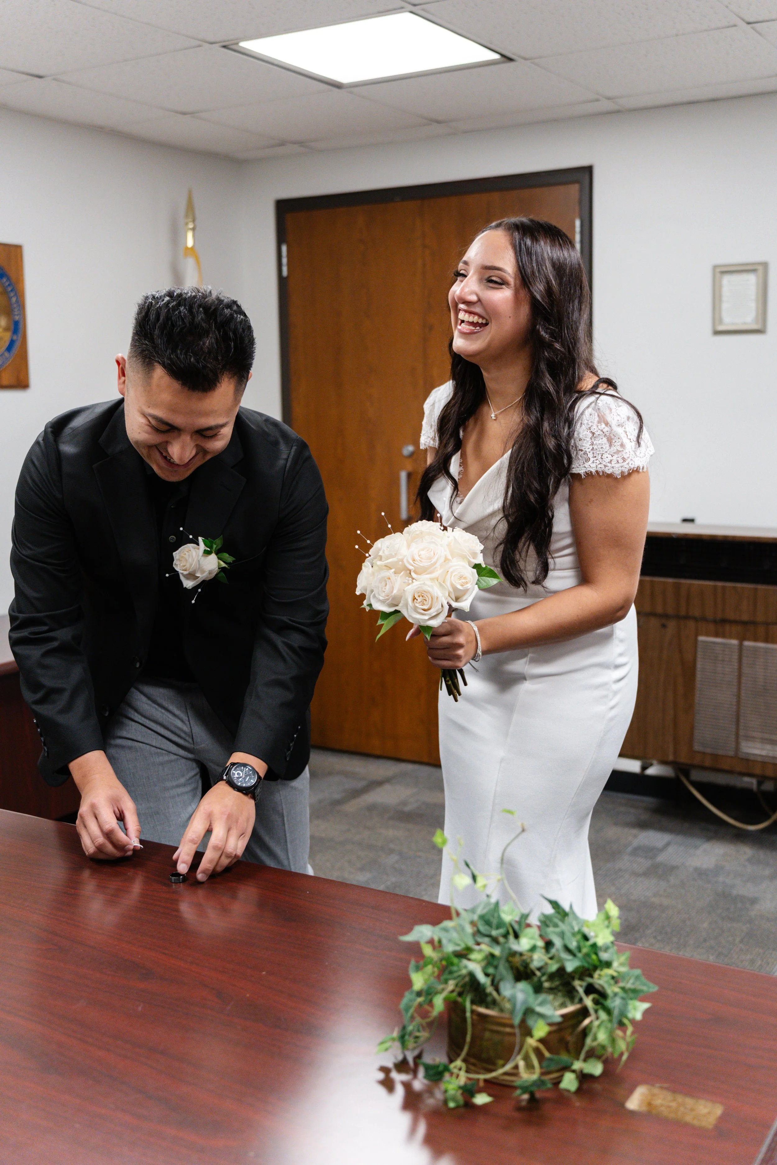 A woman in a white dress holding a bouquet of cream roses and a man in a black suit bending over a table, doingsomething on it, both smiling inside a room with brown door and wood furniture.