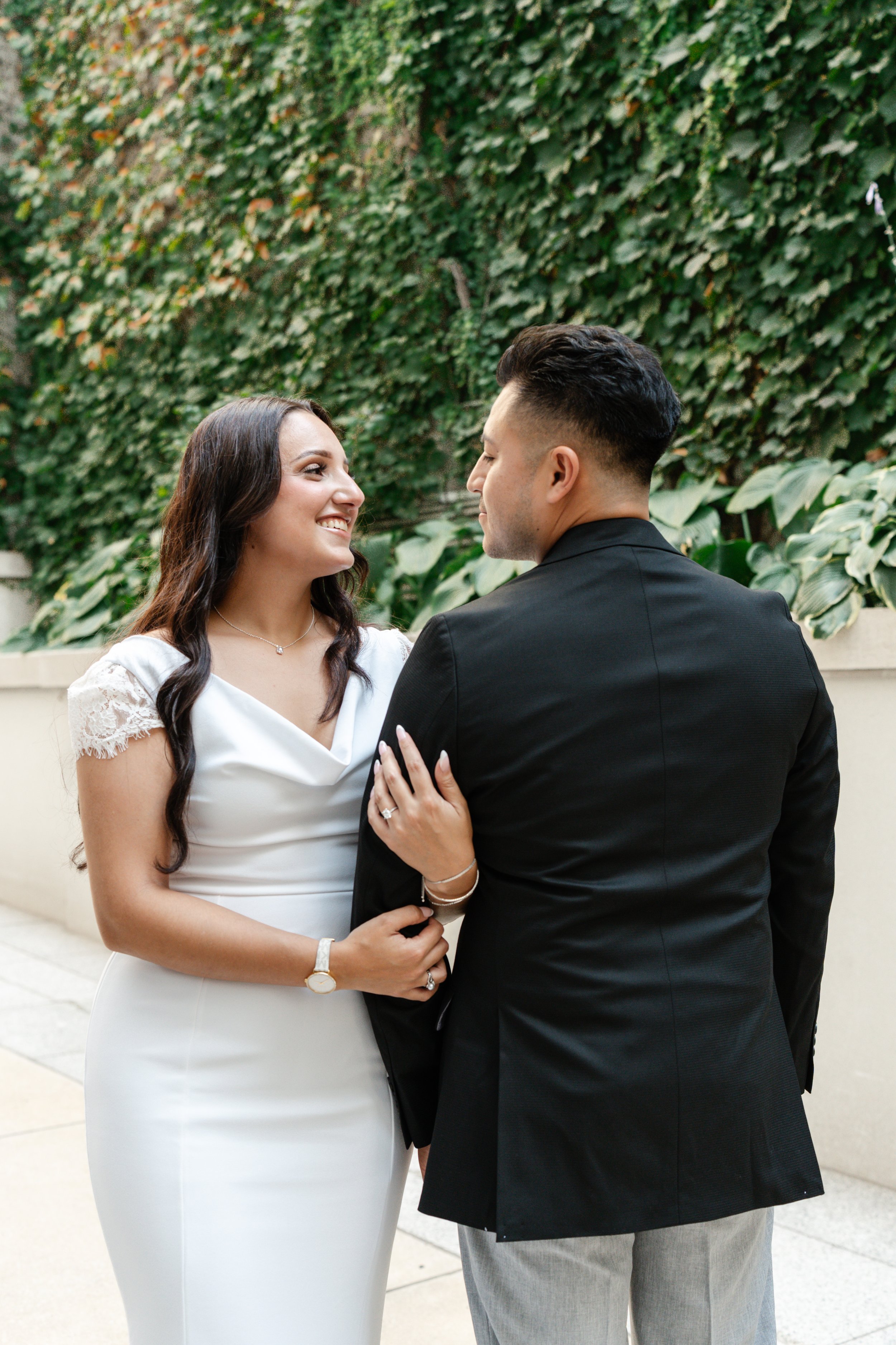 A bride and groom facing each other, smiling, standing outdoors in front of a lush ivy-covered wall, holding hands.
