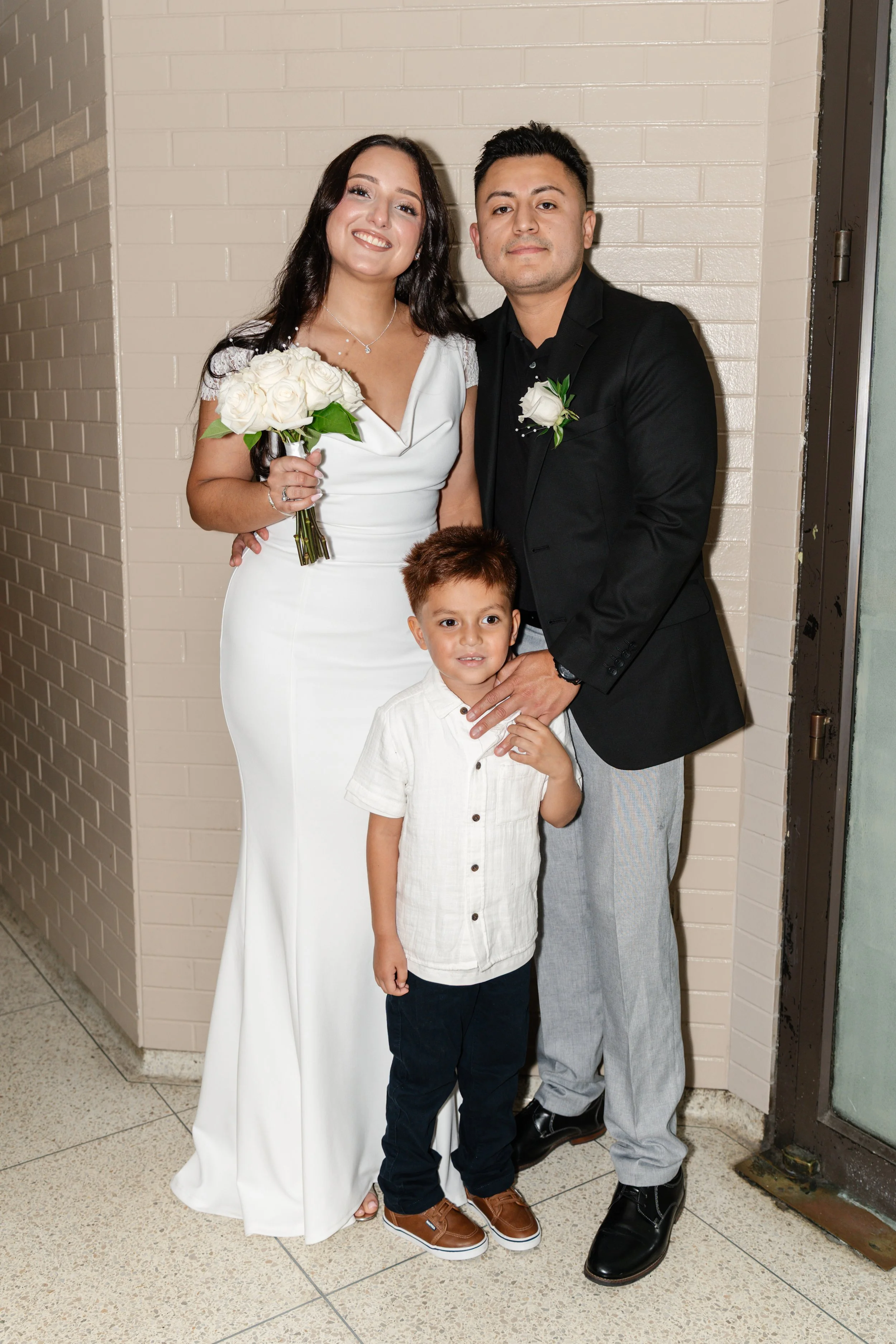 A family of three standing together indoors, with the woman in a white dress holding a bouquet of white roses, the man in a black blazer and a boutonniere, and a young boy in a white shirt and dark pants.