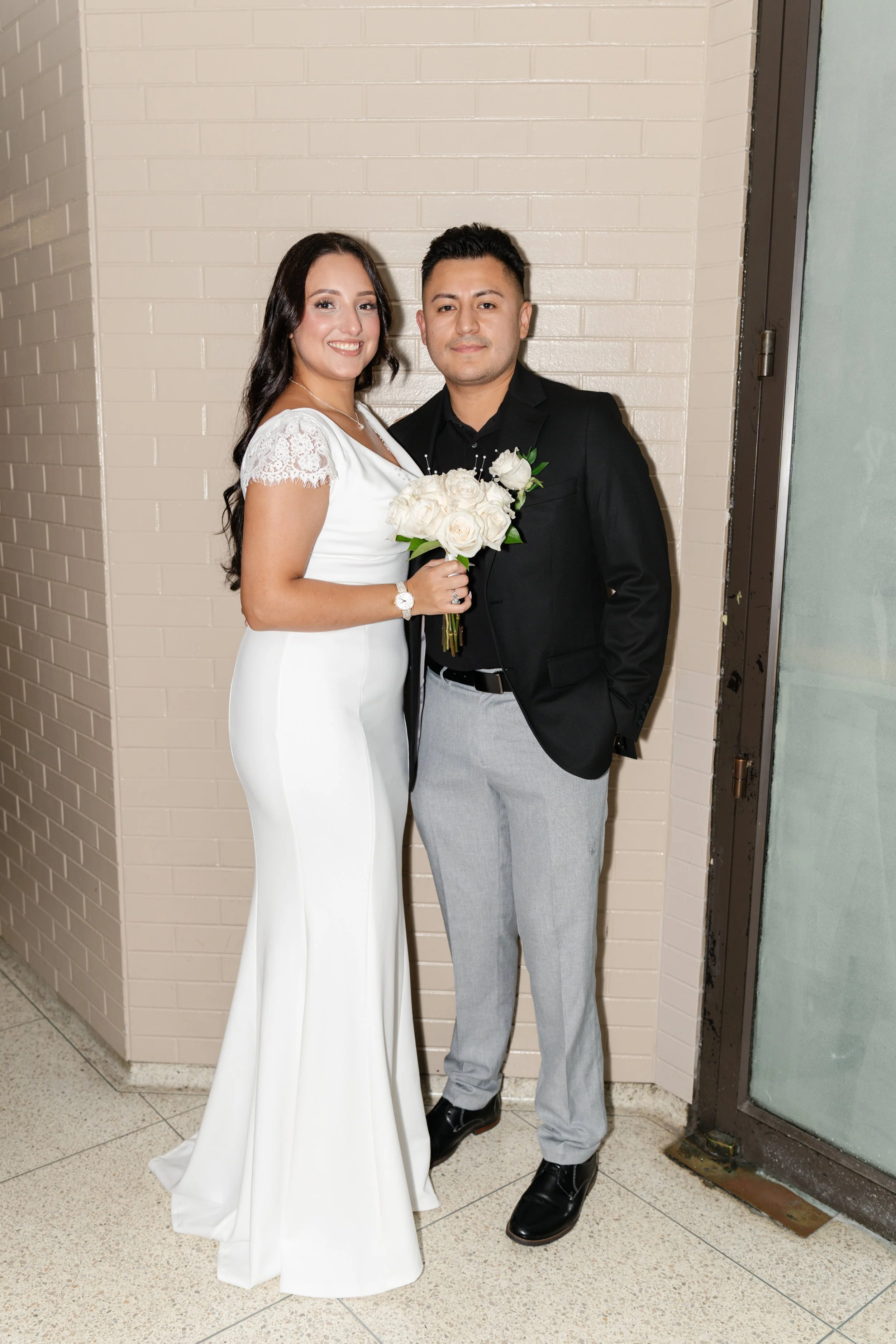 A couple dressed in wedding attire standing next to each other against a beige brick wall, with the woman holding a bouquet of white roses.