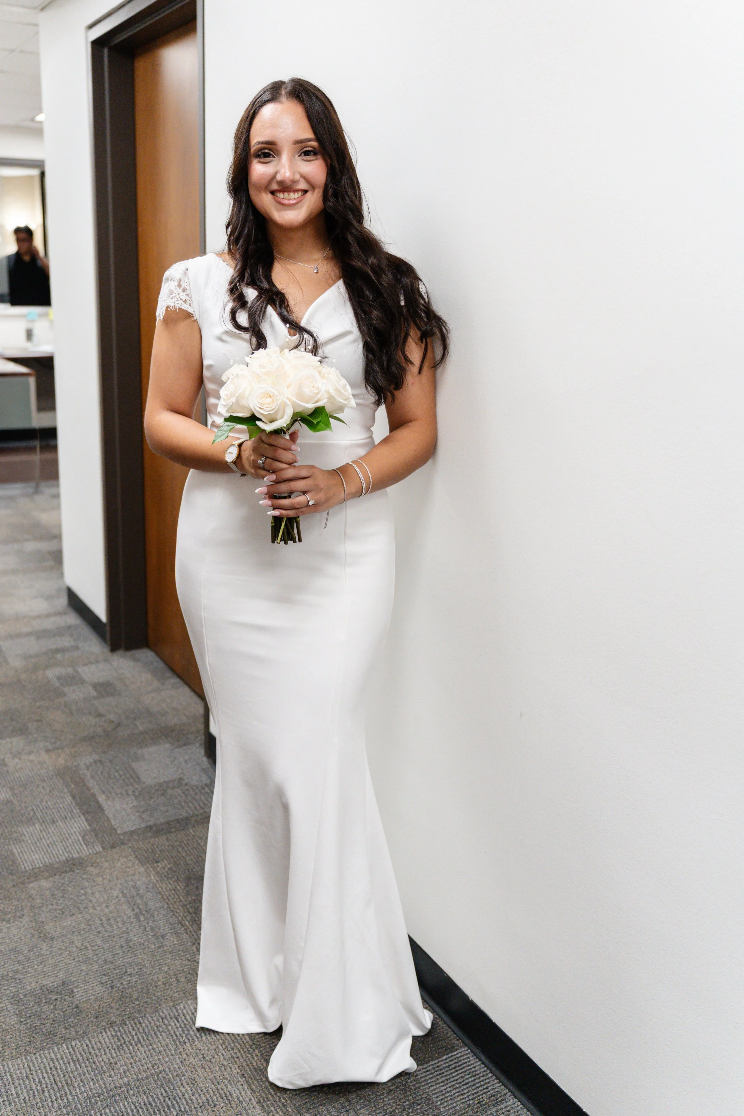 A woman in a white satin dress holding a bouquet of white roses, standing indoors near a white wall, smiling.