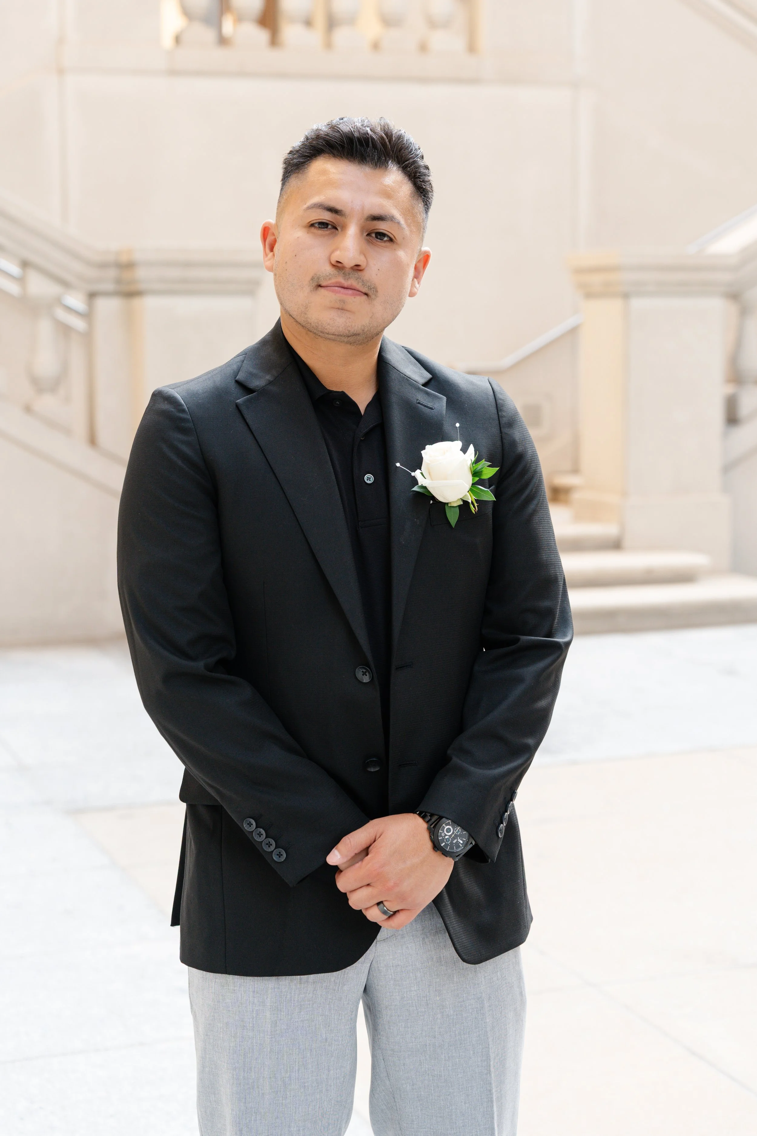 A man dressed in formal attire standing on a stone staircase, wearing a black suit jacket with a white boutonniere, a black shirt, and gray pants.