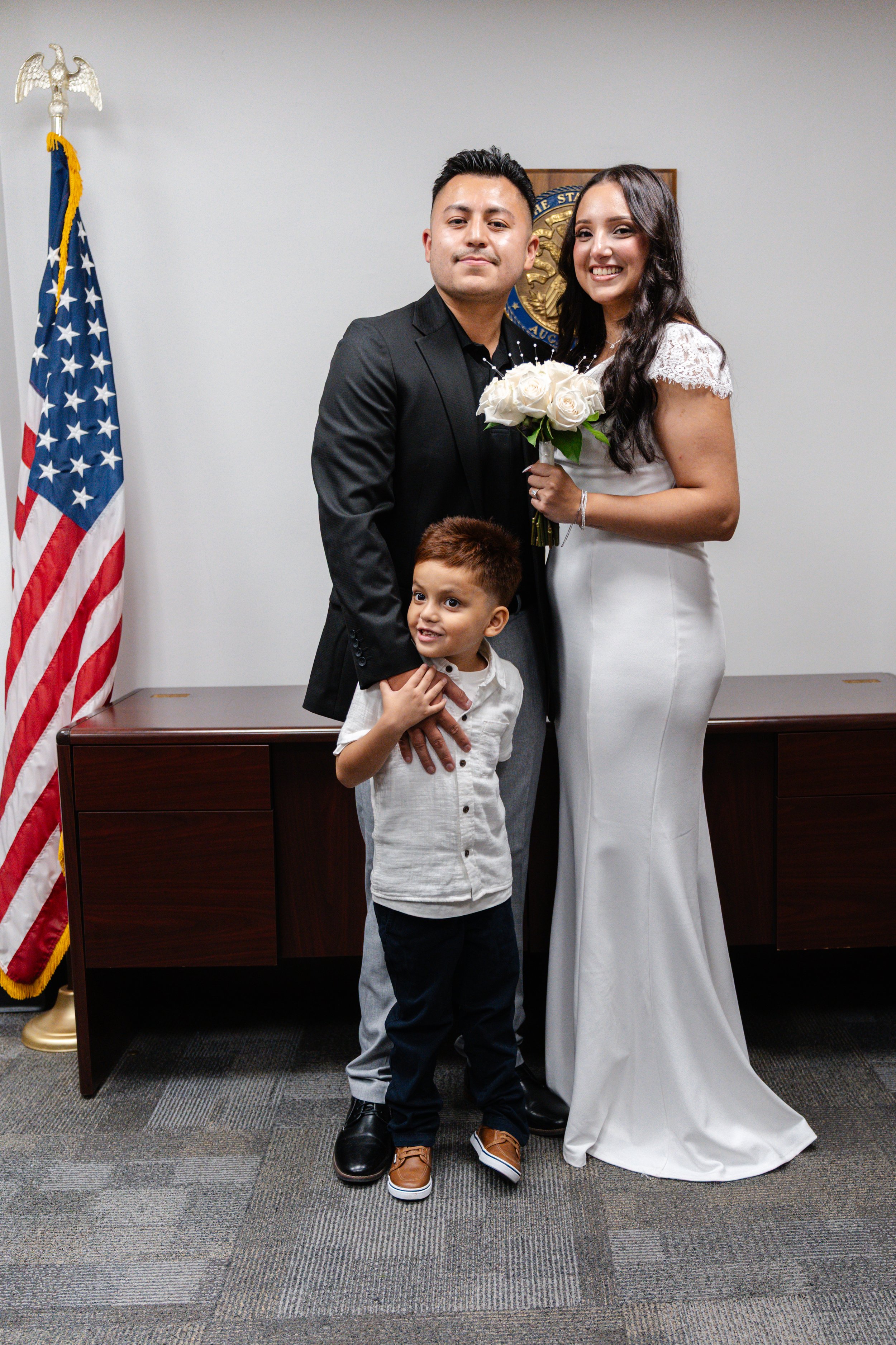 A couple with a young boy, standing in a room with an American flag and a government emblem. The woman is holding a bouquet of white roses, and all are smiling at the camera.