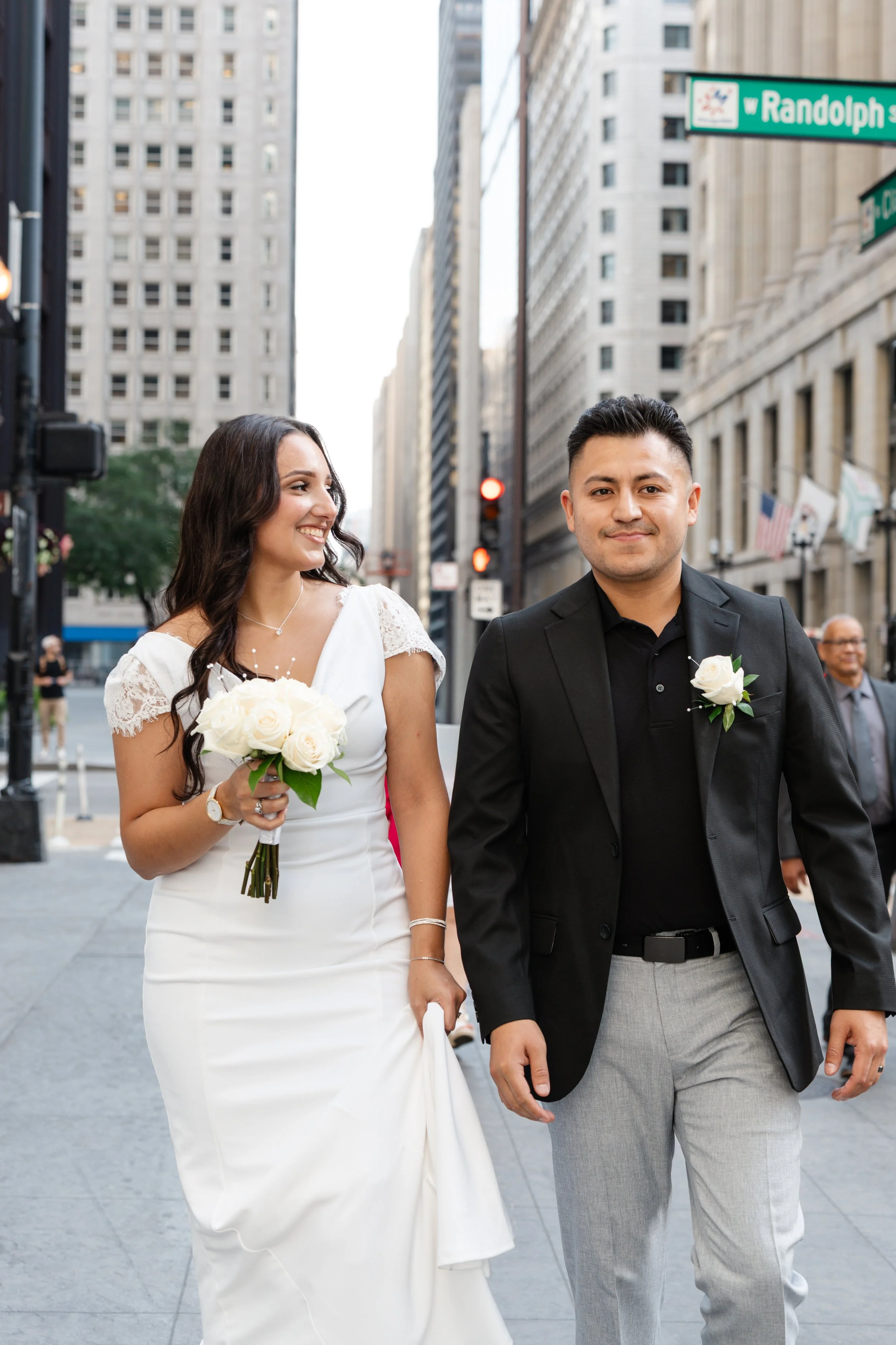 A newlywed couple walking on a city street in front of tall buildings. The bride is holding a bouquet of white roses and looking at her groom with a smile, while the groom is dressed in a black jacket, black shirt, and light gray trousers, smiling at