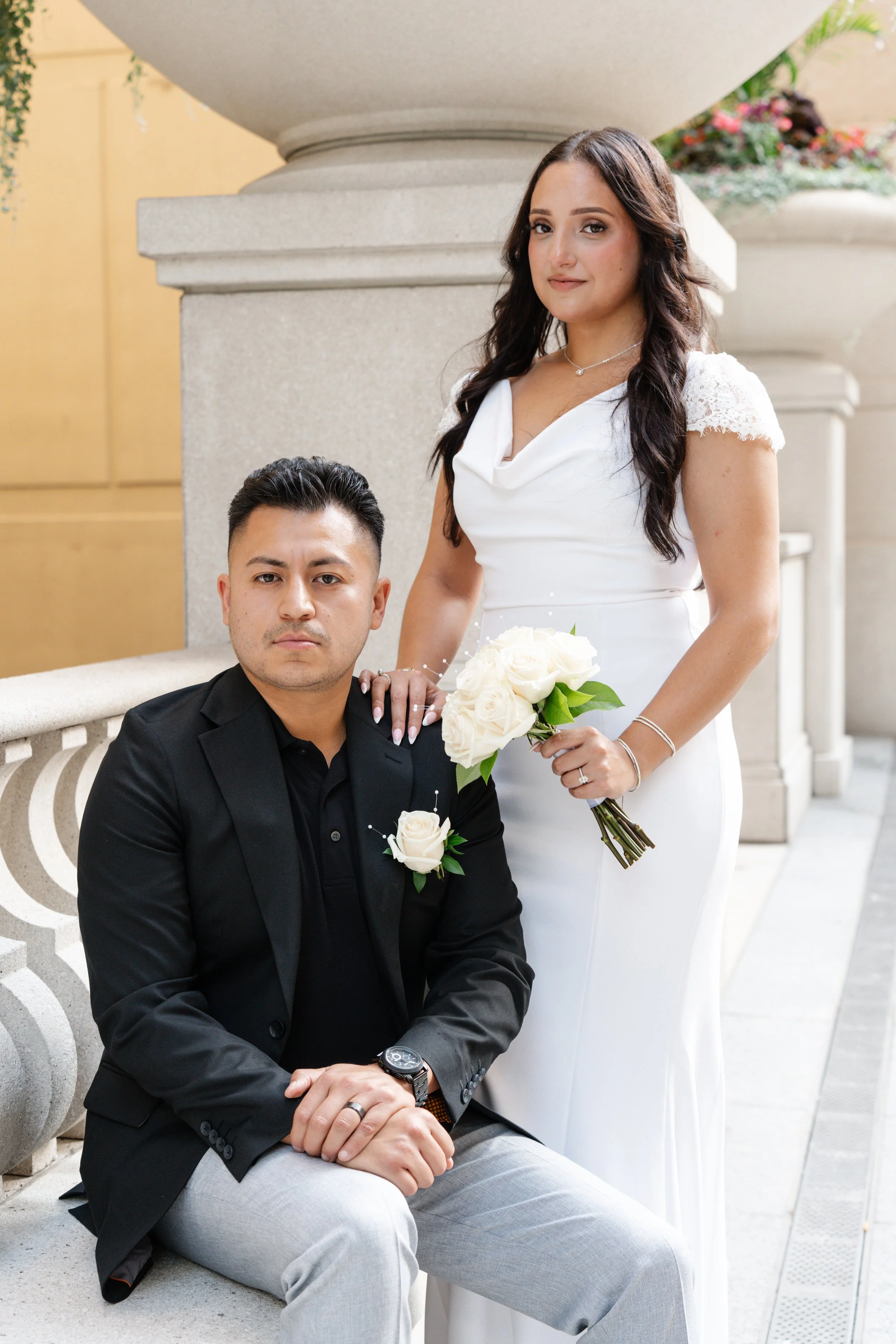 A bride in a white dress holding a bouquet of white roses stands beside a groom in a black blazer and gray pants who is seated on a stone bench, in front of a large stone balustrade and urn planters with flowers.