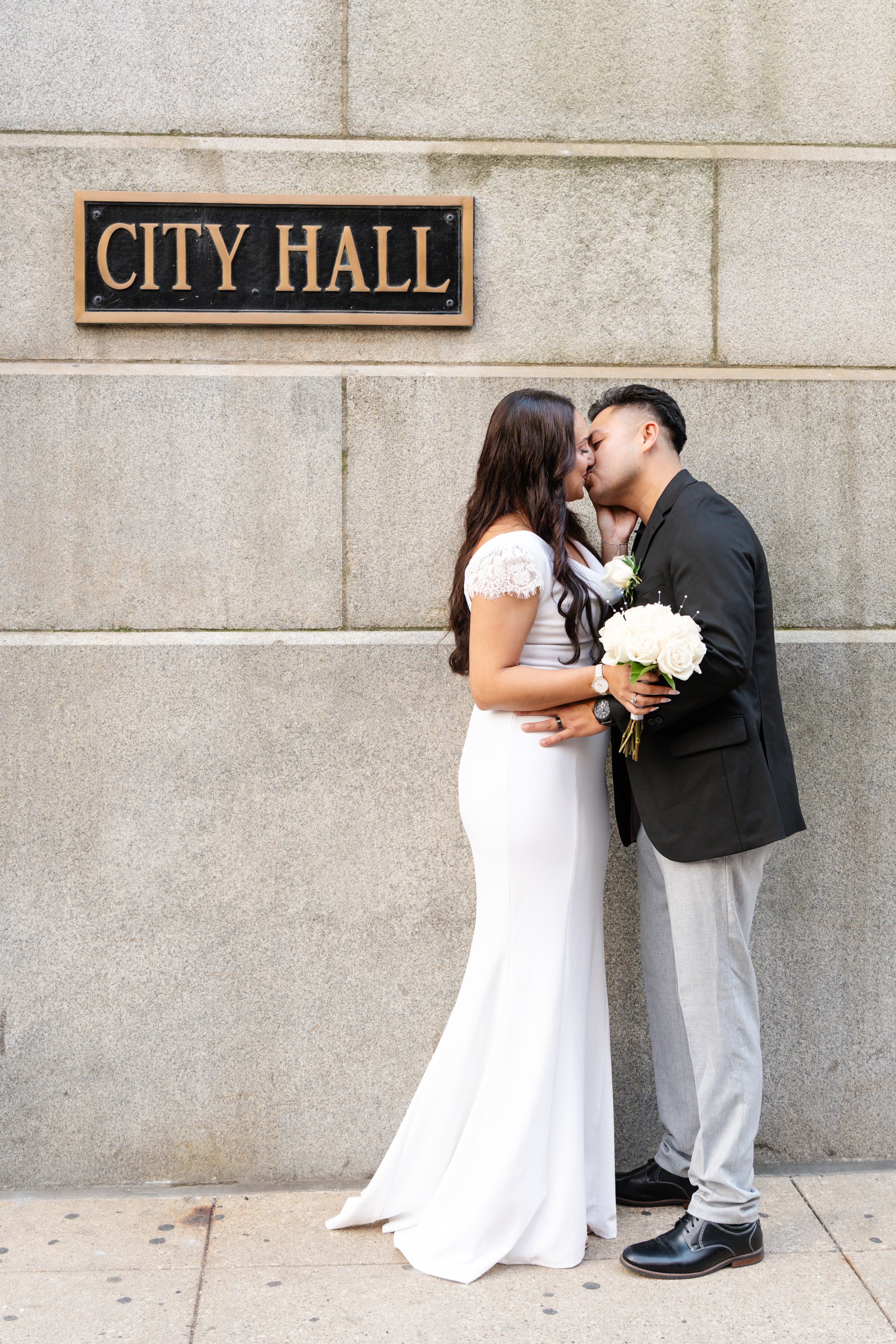 A couple dressed in wedding attire sharing a kiss outside a building with a "City Hall" sign.