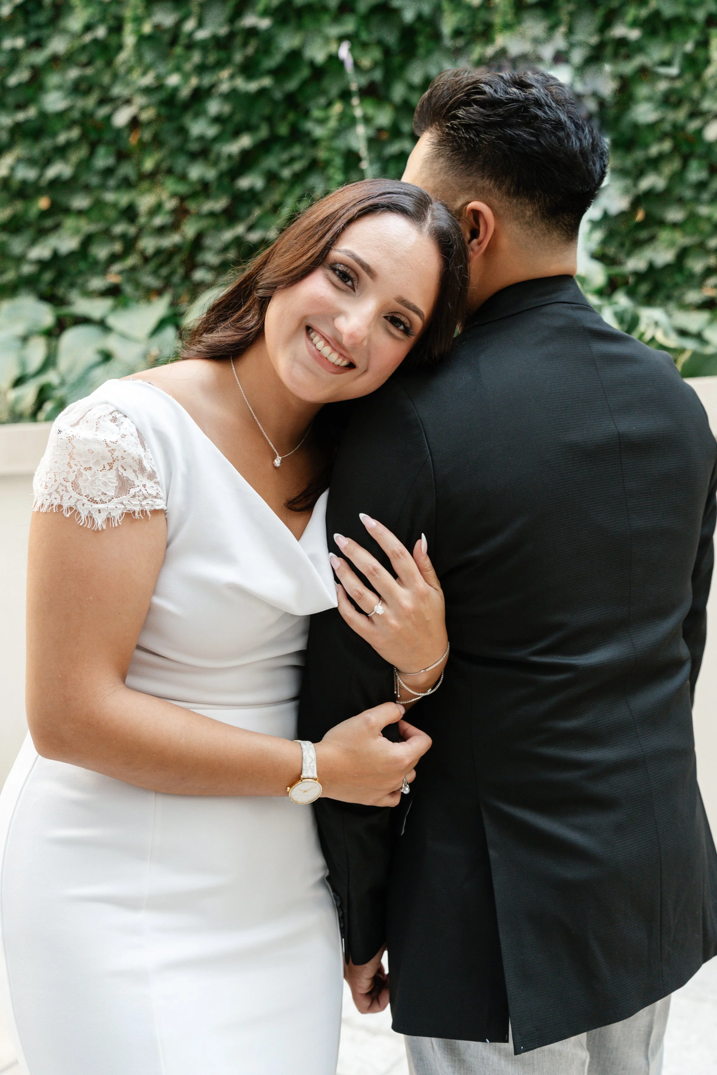 A smiling woman in a white dress with lace sleeves embraces a man in a black suit with her head resting on his shoulder, outdoor setting with greenery in the background.