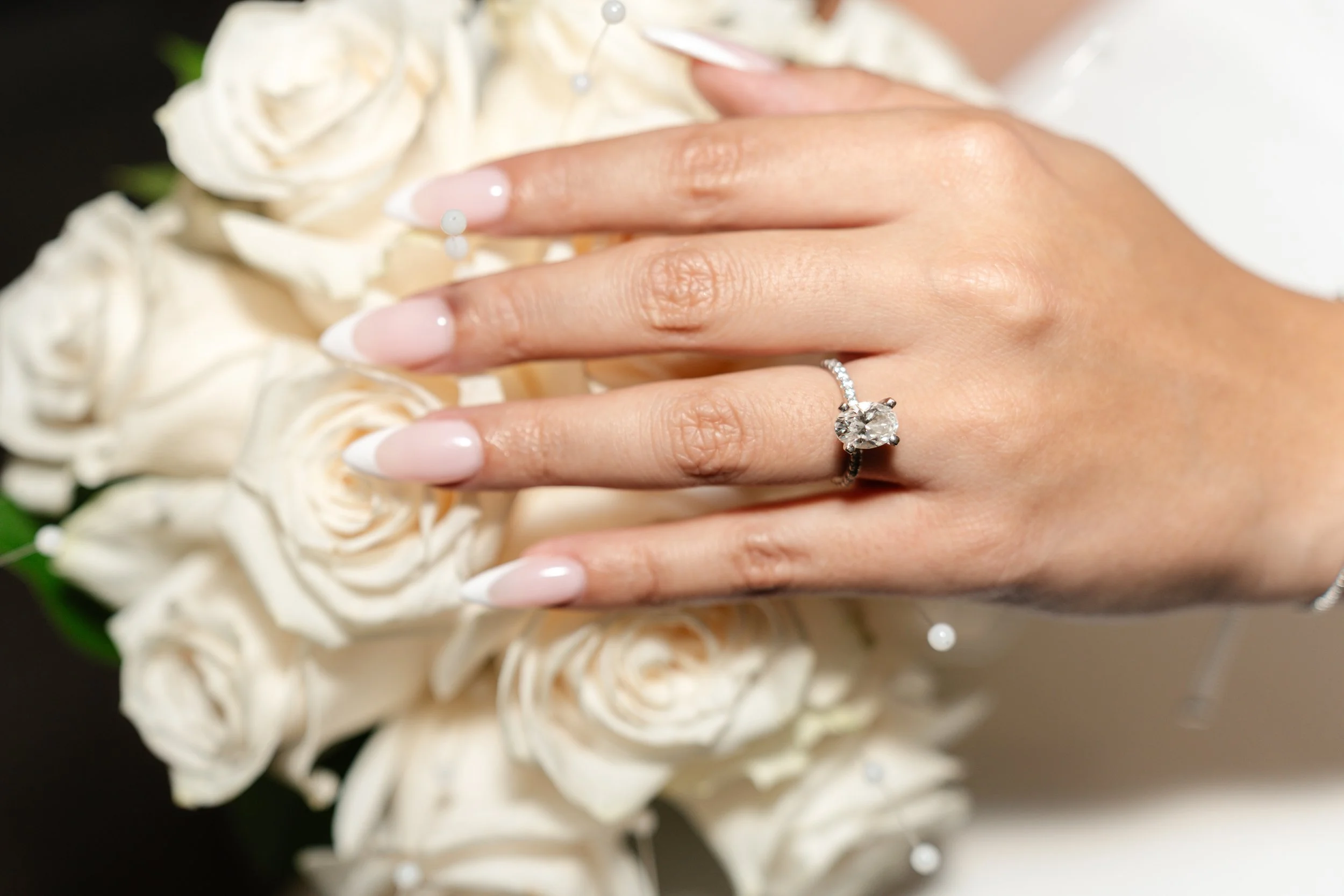 Close-up of a woman's hand wearing a diamond engagement ring, resting on white roses.