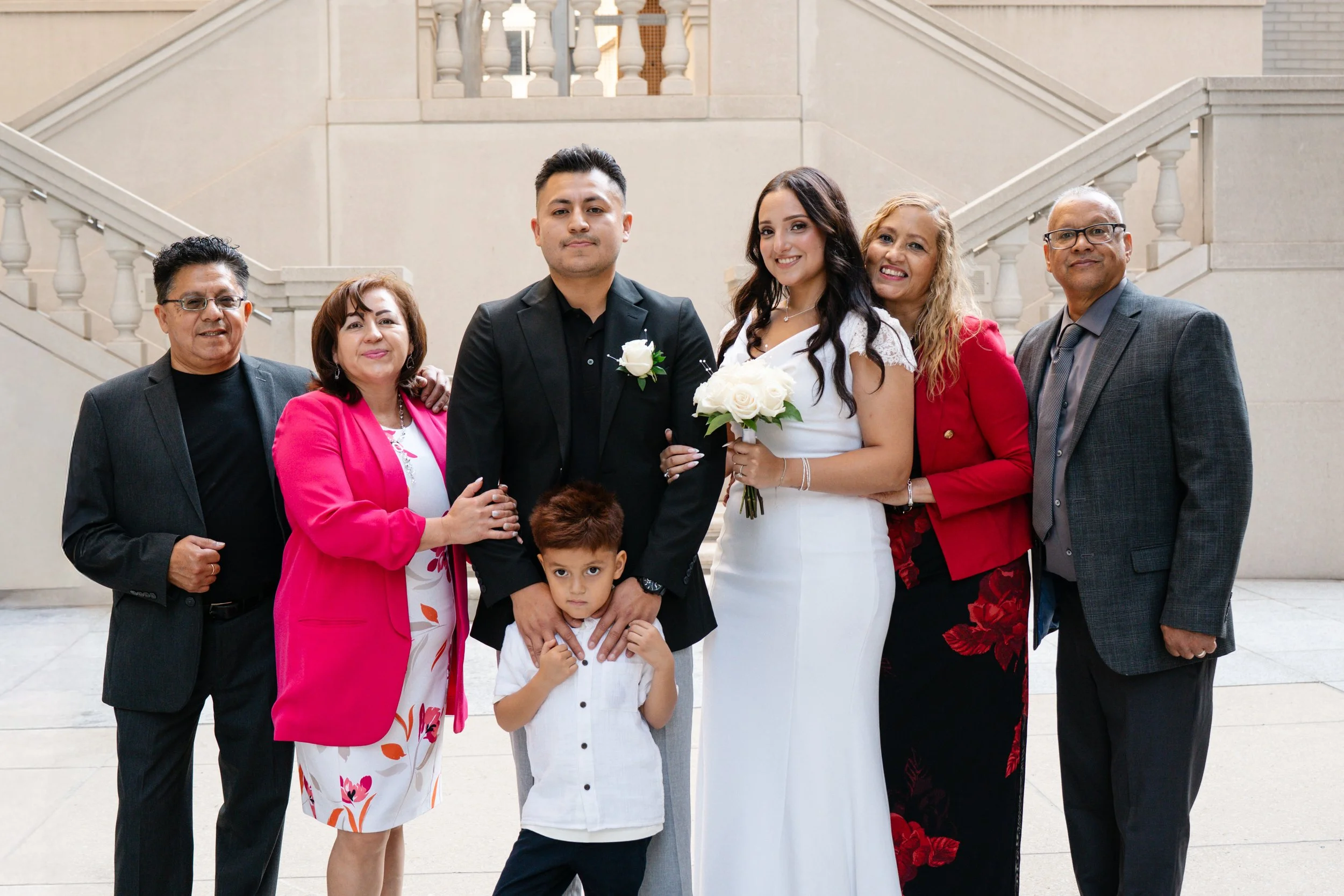 Family group photo at a wedding, with bride and groom in the center, holding flowers, surrounded by family members dressed in formal attire.