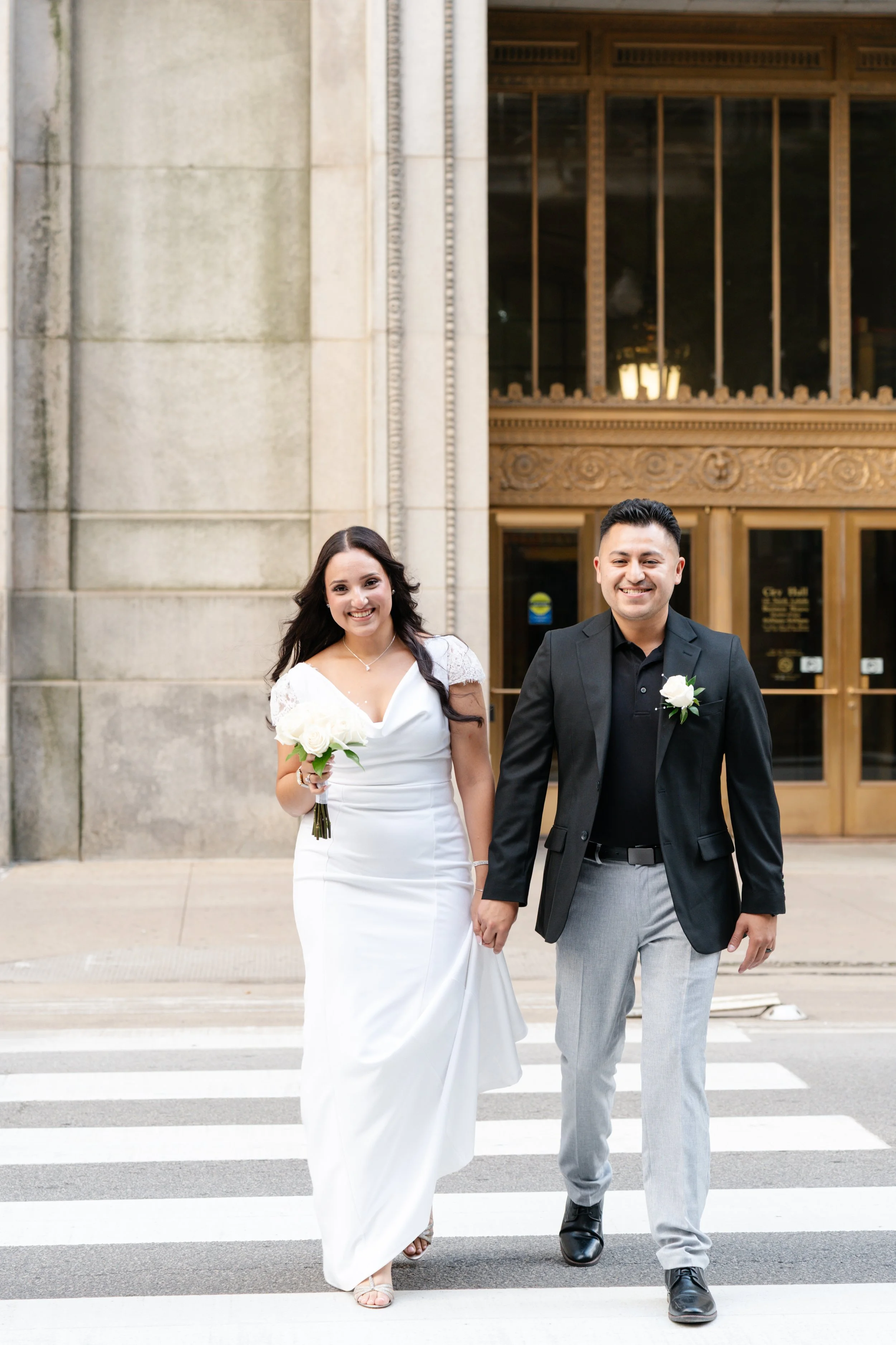 A newlywed couple walking across a crosswalk outside a building, holding hands and smiling, with the bride wearing a white wedding gown and holding a bouquet, and the groom dressed in a dark blazer and light gray pants.