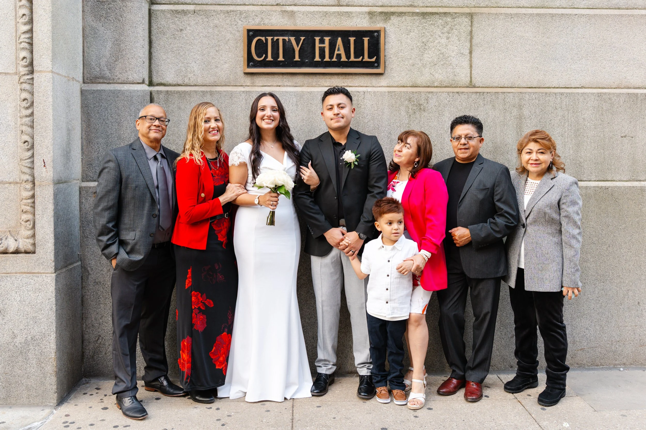 A diverse group of nine people, including a bride and groom, posing in front of a city hall sign. The bride is in a white dress holding a bouquet, and the groom is in a black jacket with a boutonniere. The group is dressed formally, with some women i