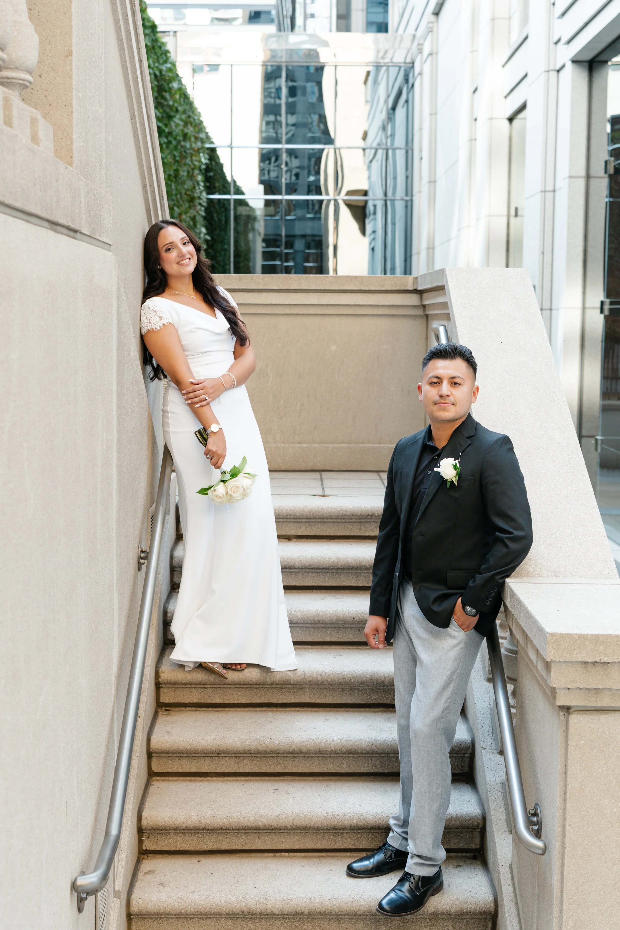 A couple dressed in formal attire on outdoor stairs in an urban setting. The woman is wearing a white dress and holding a small bouquet of white roses, while the man is in a black blazer with a white rose boutonniere and gray trousers.