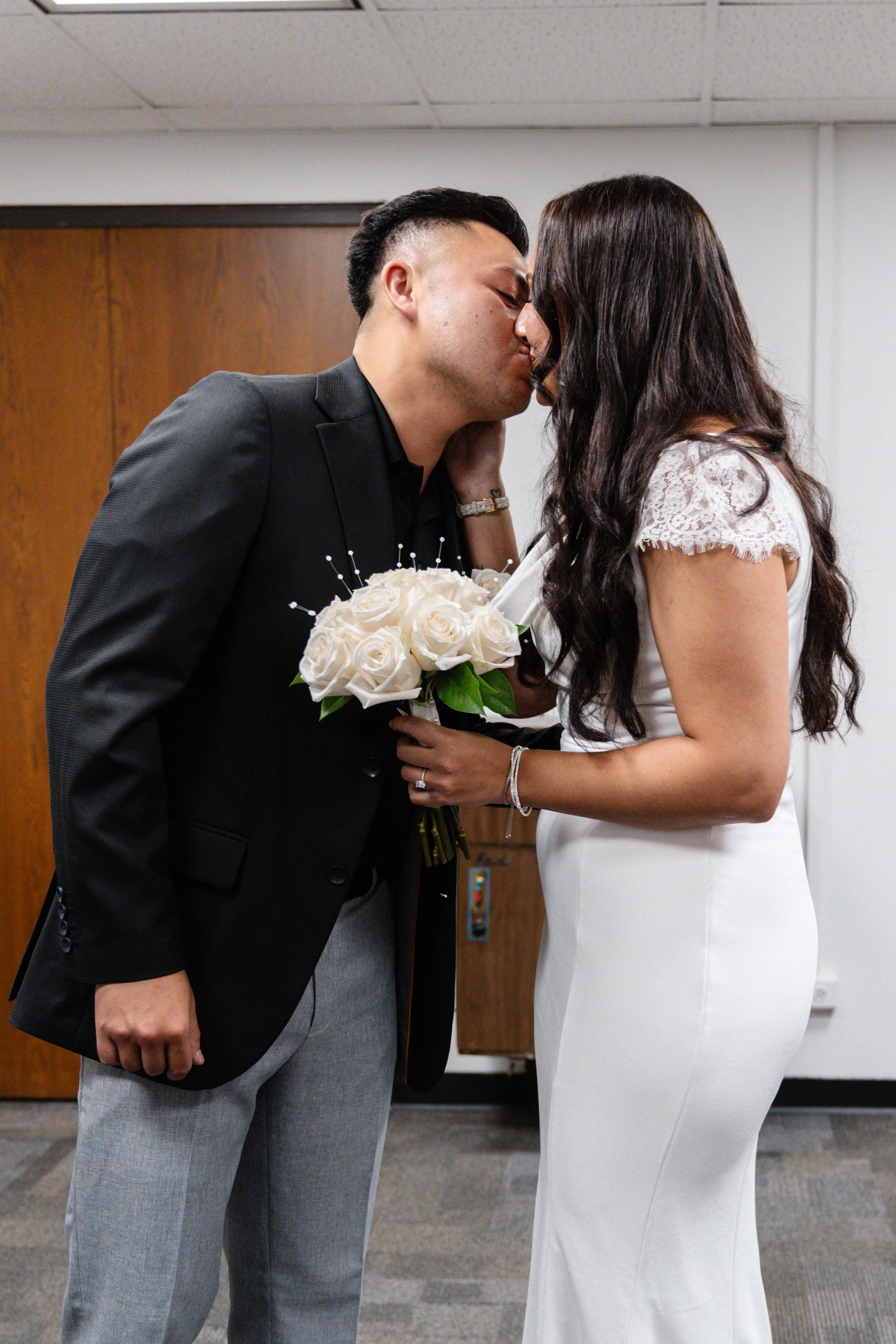 A couple in wedding attire sharing a kiss with the bride holding a bouquet of white roses.