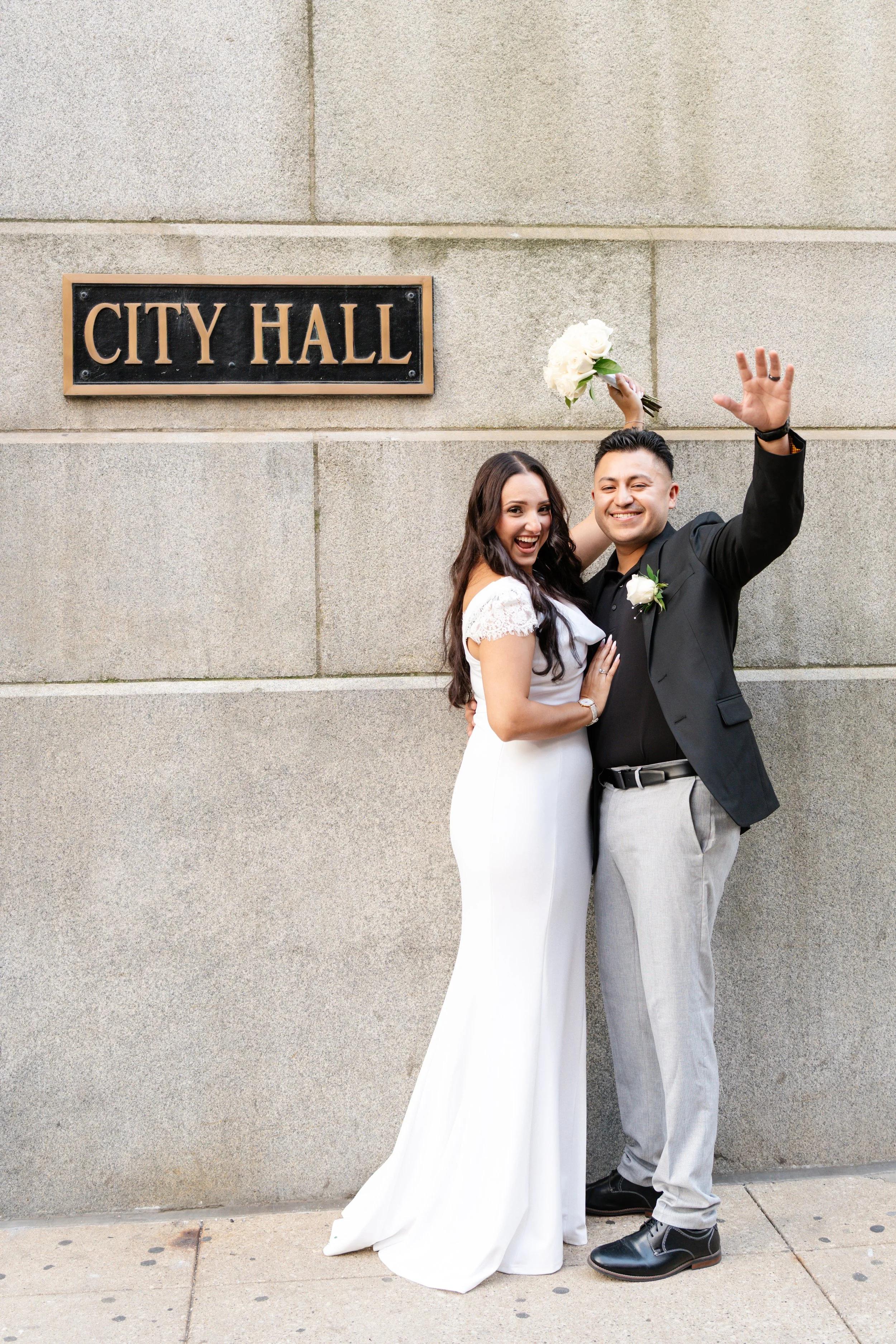 A newly married couple celebrating outside a city hall building. The bride is in a white wedding dress, holding a bouquet, and smiling. The groom is in a black blazer, gray pants, and is waving with a big smile. There is a "City Hall" sign on the wal