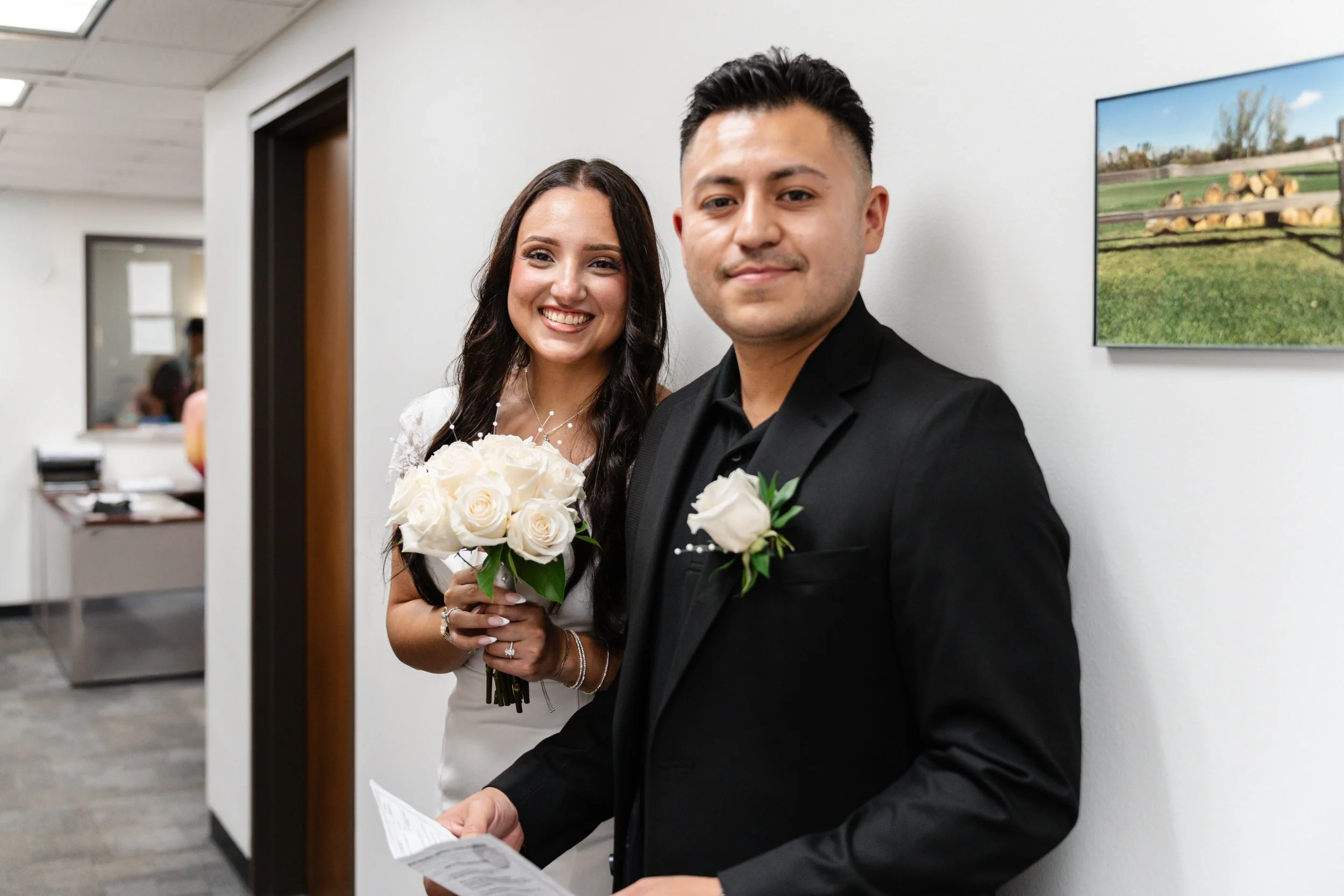 A newlywed couple stands in a hallway, smiling for a photo. The bride holds a bouquet of white roses, and both wear white flowers pinned on their outfits. The groom is dressed in a black suit, and the bride in a white dress.